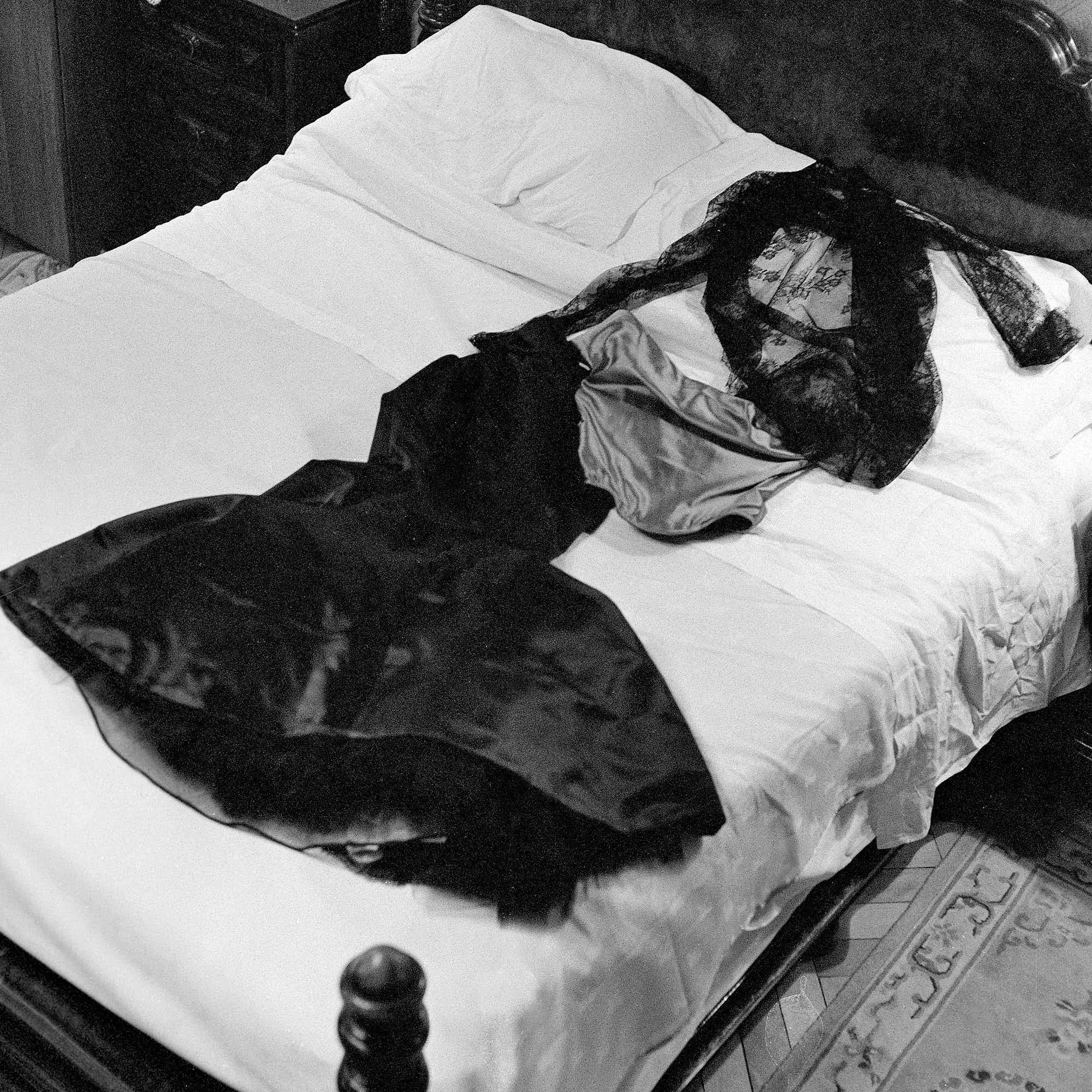 A black and white photo of a bed with white sheets, a pillow, and a lace curtain draped over the side, in front of a dark headboard.
