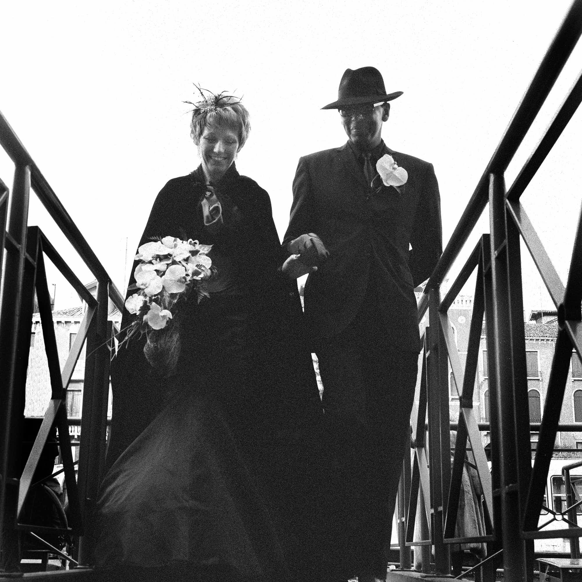 Black and white photo of a man and woman holding hands, walking up stairs. The woman is holding a bouquet of flowers and wearing a hat with decorative feathers. The man is wearing a fedora and eyeglasses, with a flower pinned on his lapel. They are s