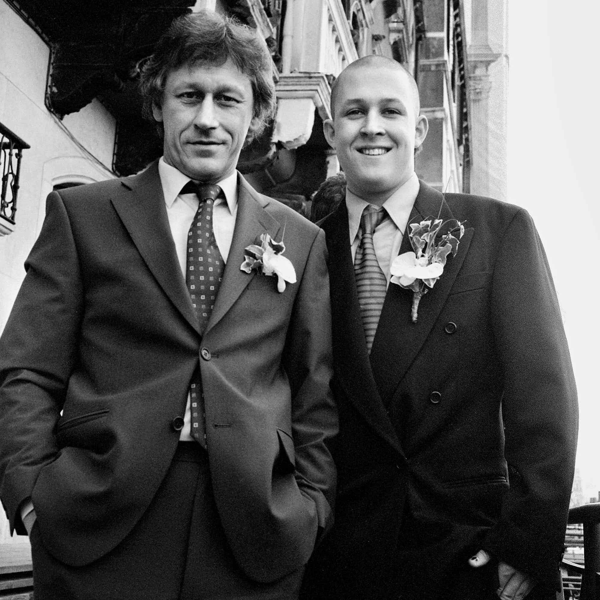 Two men in suits standing close together outdoors; one has wavy hair and the other is bald, both smiling, with a backdrop of a building with a fire escape and some trees.