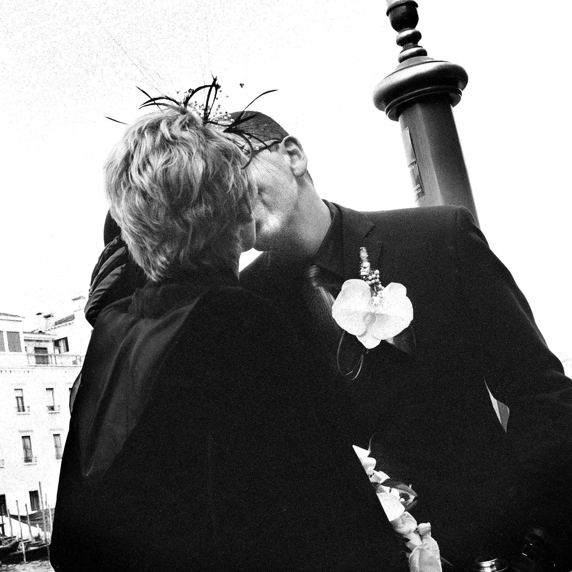 A black and white photo of a person with short, light-colored hair standing outdoors next to a lamp post. They are wearing a dark outfit and holding a bunch of flowers, leaning in for a kiss with another person whose face is not visible. Buildings ar