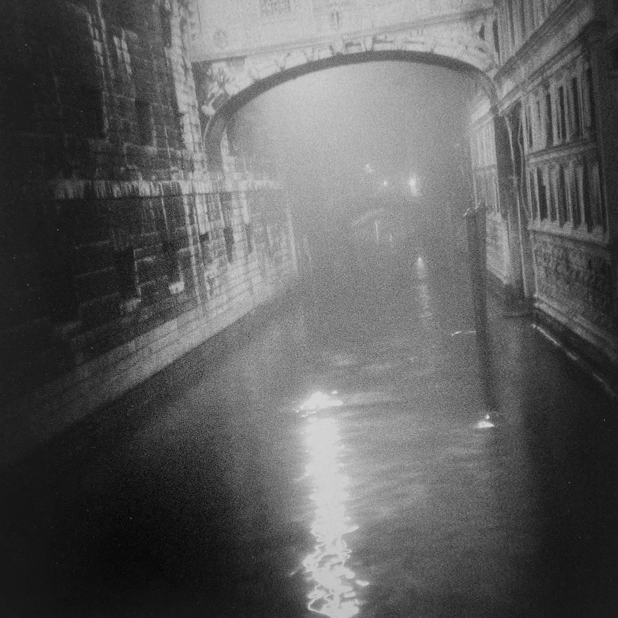Nighttime scene of reflections on wet cobblestone street with an arched bridge and old buildings lining the street.