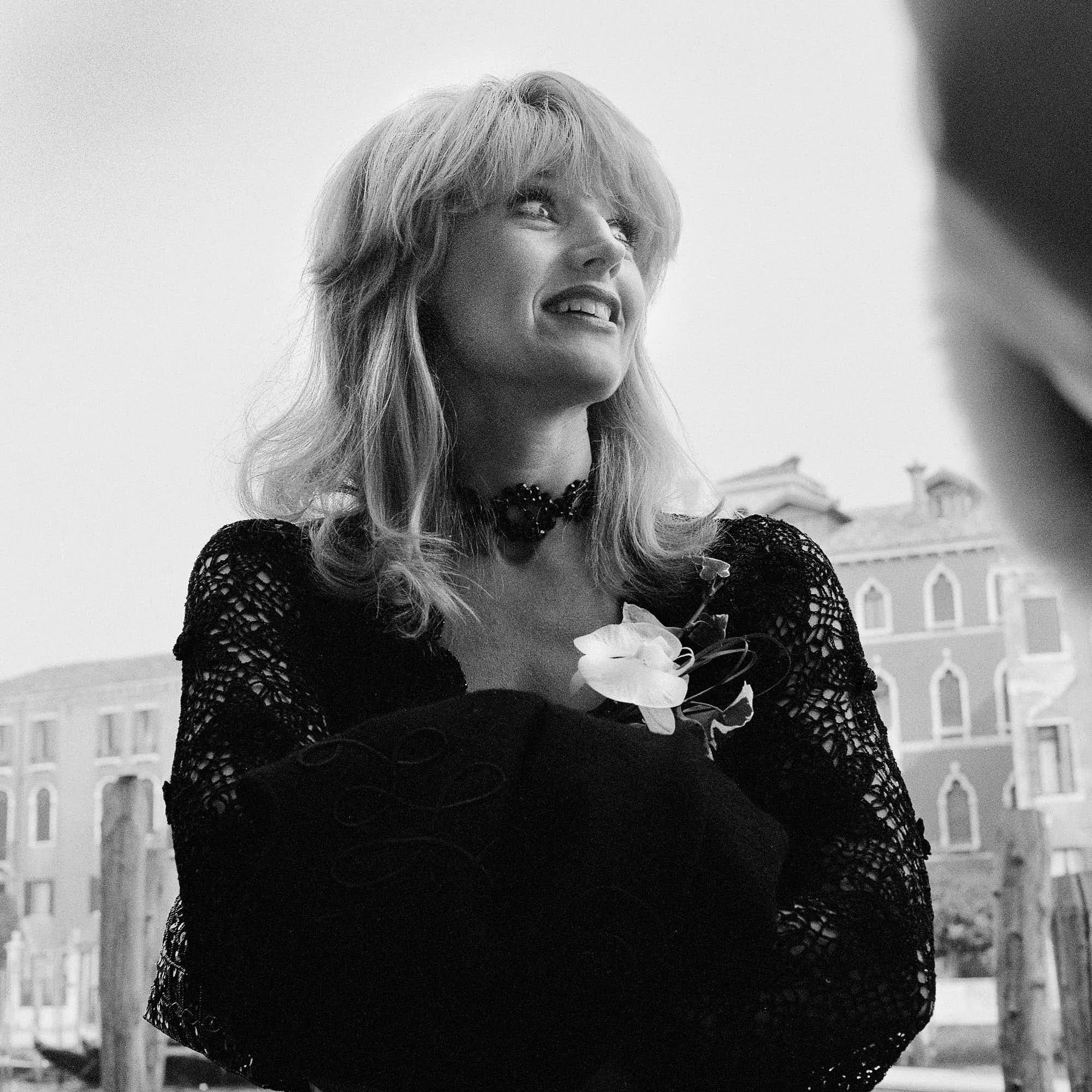 Black and white photo of a woman with blonde hair, smiling and holding flowers, standing outdoors with buildings in the background.