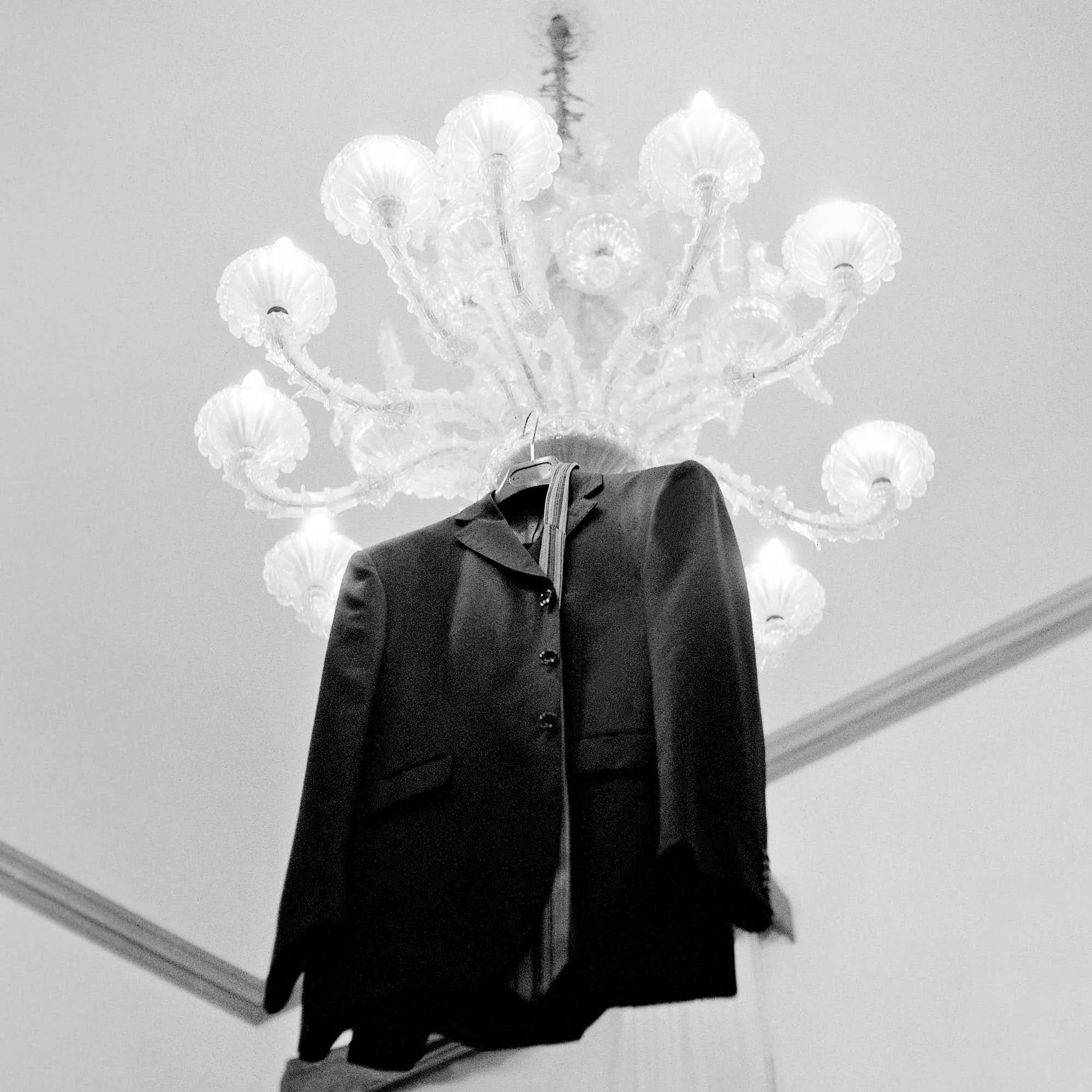A person in a suit taking a photo in a mirror, with a chandelier hanging from the ceiling in the background.