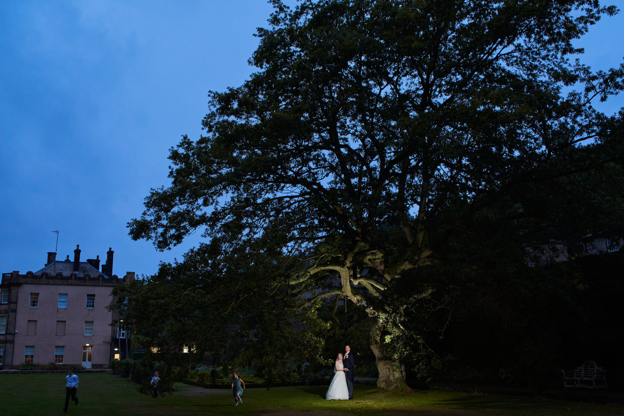 A bride and groom standing under a large tree at dusk, with children playing nearby in an outdoor garden setting. Buildings are visible in the background.