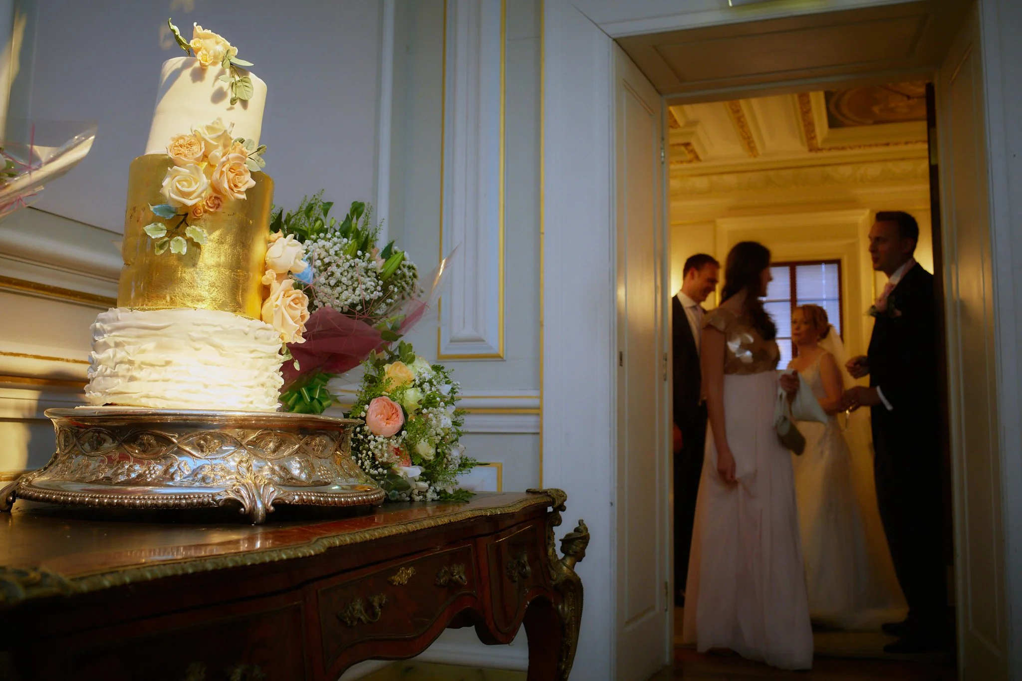 A wedding cake decorated with white fondant, peach and white roses, and green leaves, sitting on an ornate silver cake stand. In the background, a couple and two women are standing and talking inside a richly decorated room with ornate ceiling detail