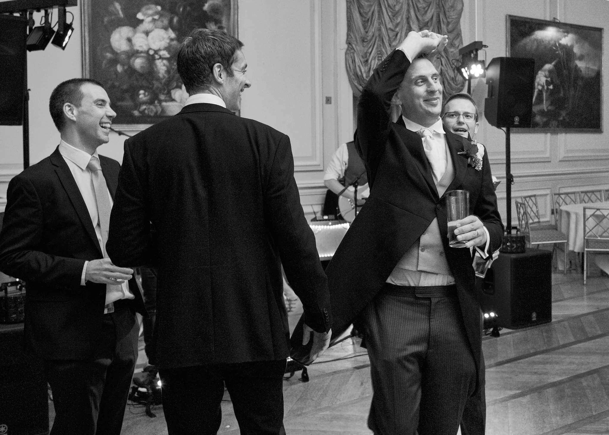 Four men dressed in tuxedos at a wedding reception. They are smiling, laughing, and enjoying the celebration. One man is holding a drink and the others are engaging in conversation. The setting is an elegant room with paintings and drapery.