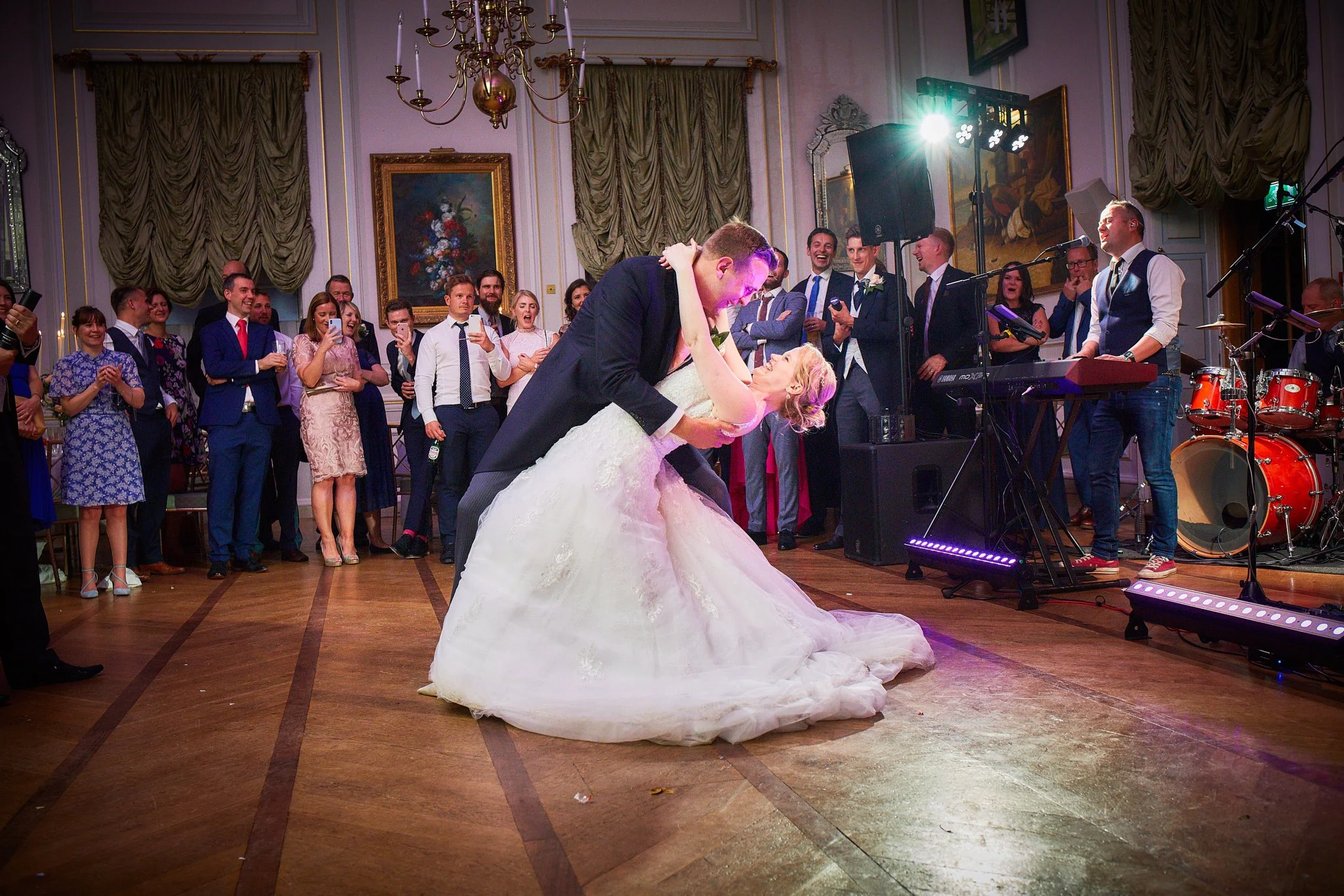 A wedding reception with a bride and groom dancing among guests, in a decorated ballroom with ornate curtains and paintings, and a live band playing music.