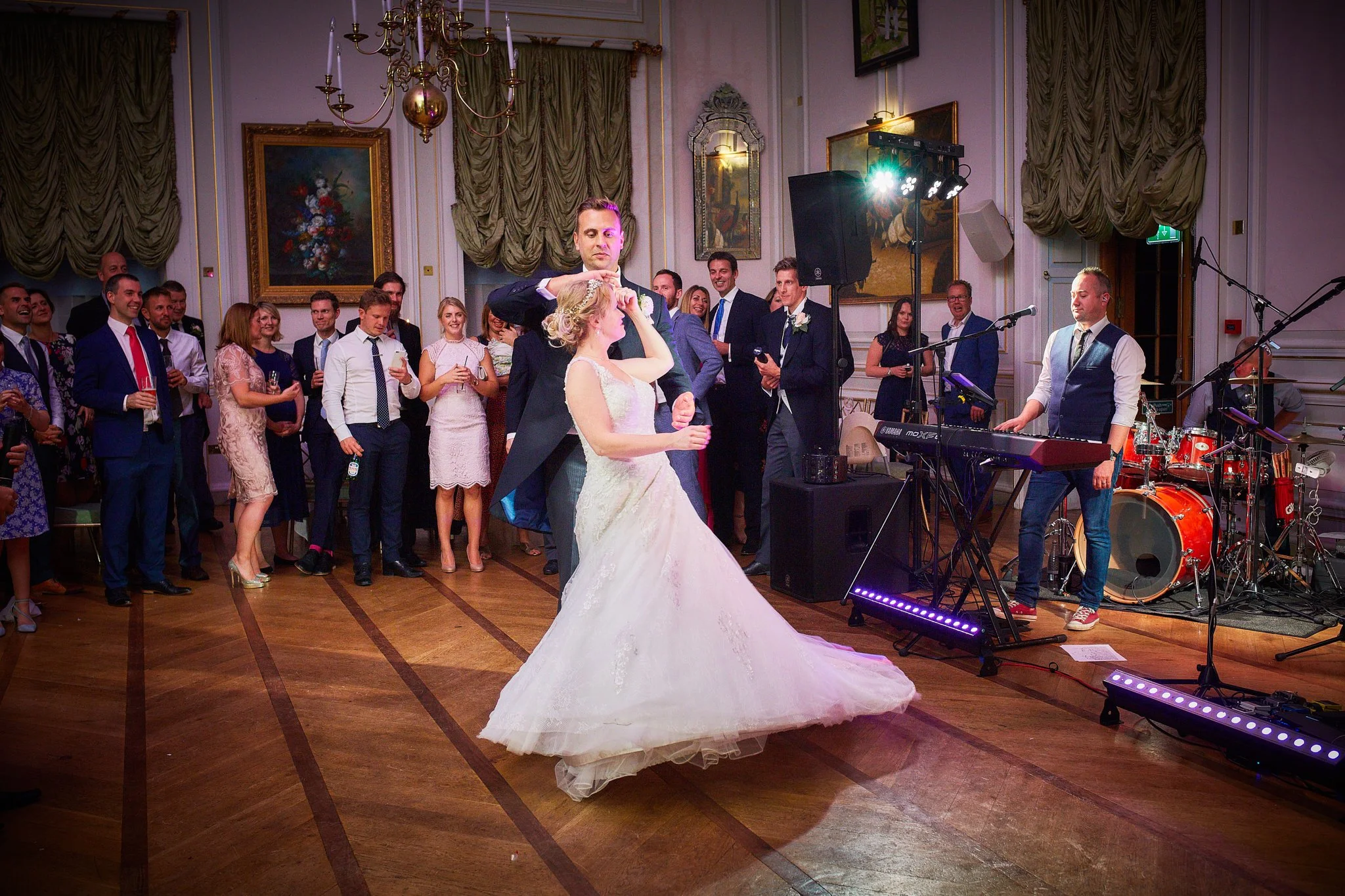 A bride and groom dancing at their wedding reception with guests watching and smiling in the background. The room has ornate decor, including paintings, curtains, and chandeliers, with a live band playing music.