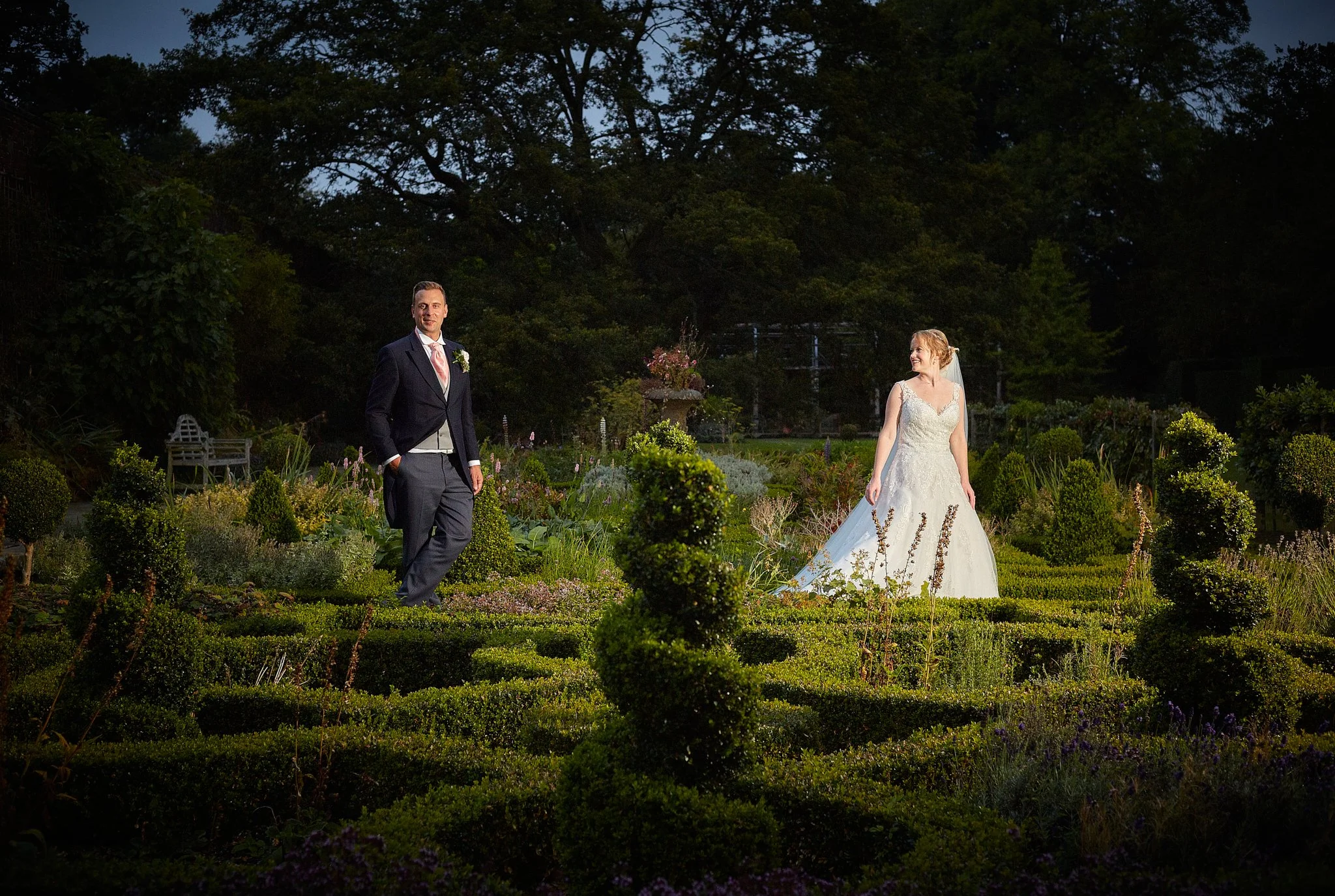 A bride and groom standing in a garden at dusk. The groom is wearing a dark suit with a pink tie and boutonniere. The bride is in a white wedding gown with lace details, looking at the groom with a smile. The garden is lush with green bushes and flow