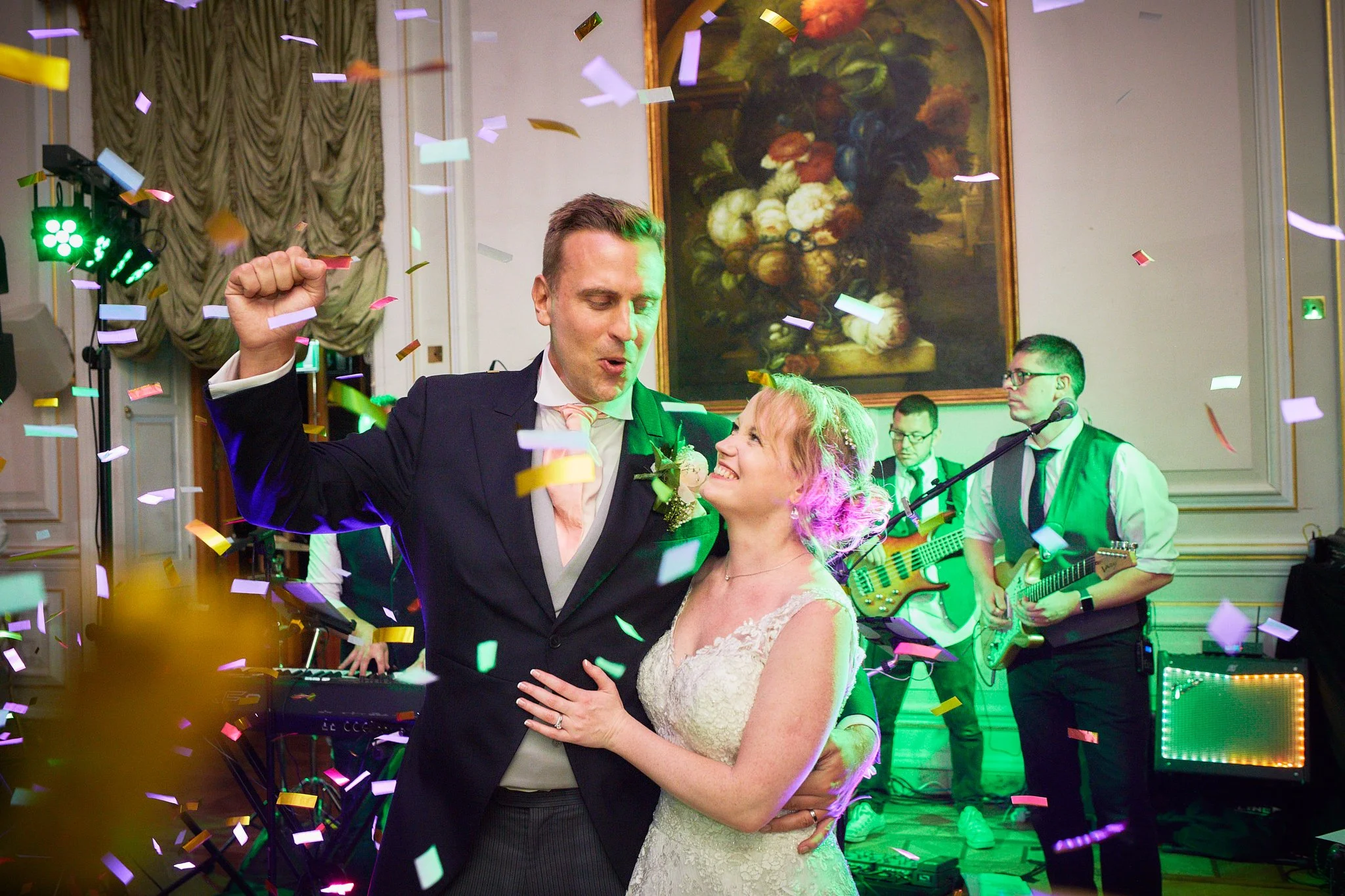 A couple dancing at a celebration with colorful confetti falling around them. The man is in a suit, and the woman is in a white lace dress. They are smiling and appear joyful. There is a band playing in the background with musicians and a large firep