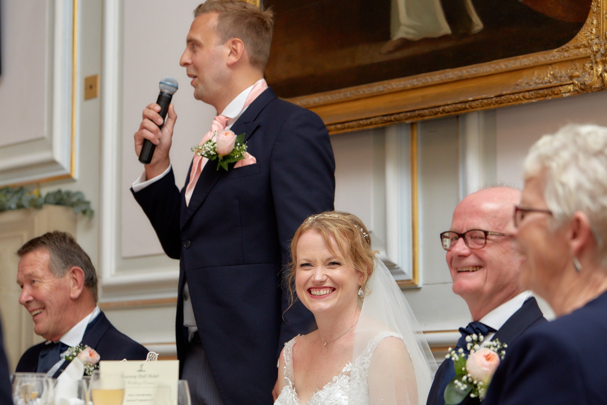 A groom giving a speech at a wedding reception, holding a microphone and wearing a dark suit with a pink tie and a boutonniere. The bride and guests are smiling and seated at a decorated table.
