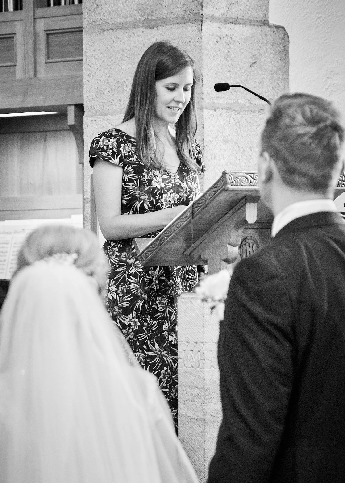 A woman is standing at a pulpit, reading or speaking, in a room with stone and textured walls. Several people are seated in front of her, some with hairstyles visible in the foreground.