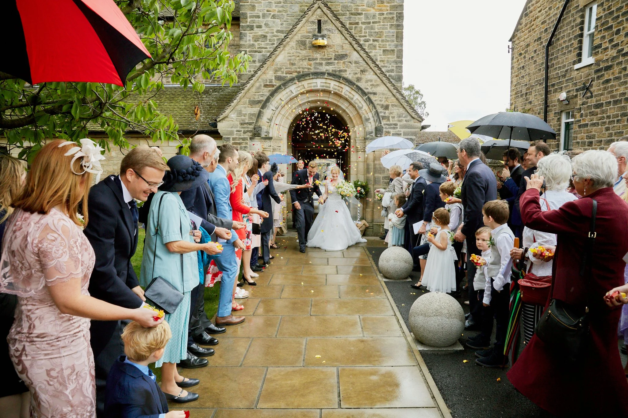 A bride and groom exit a church while guests celebrate by throwing colorful flower petals and confetti. Guests hold umbrellas due to rainy weather. The church has a stone facade and arched entrance.
