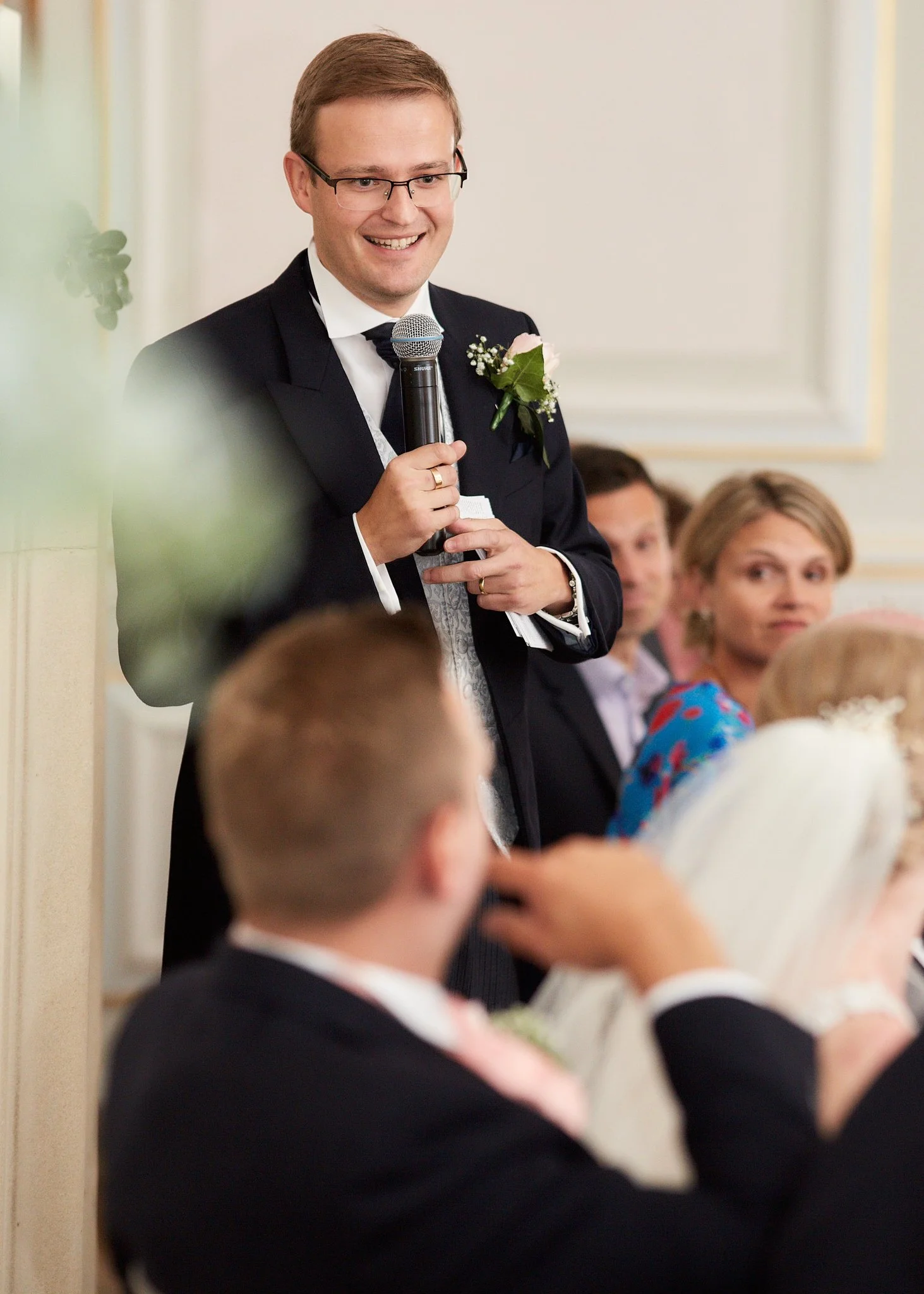 A man in a tuxedo with glasses is giving a speech at a wedding, holding a microphone and smiling. There are guests in the background, sitting and listening.