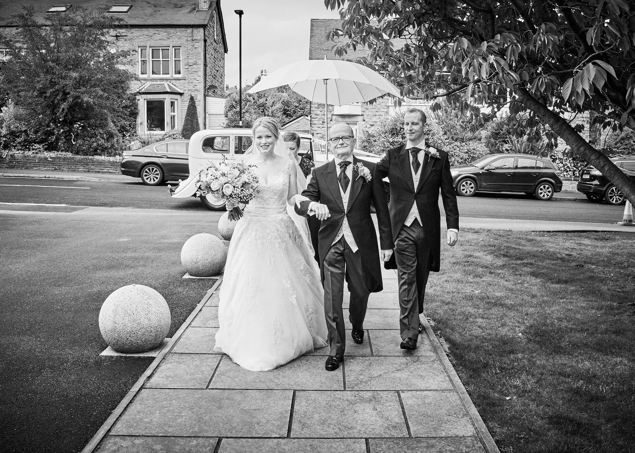 Black and white photo of a wedding procession with a bride in a wedding dress holding a bouquet, walking between two men in suits. One man holds a large umbrella over her. They are walking on a paved sidewalk with decorative stones, over a street wit