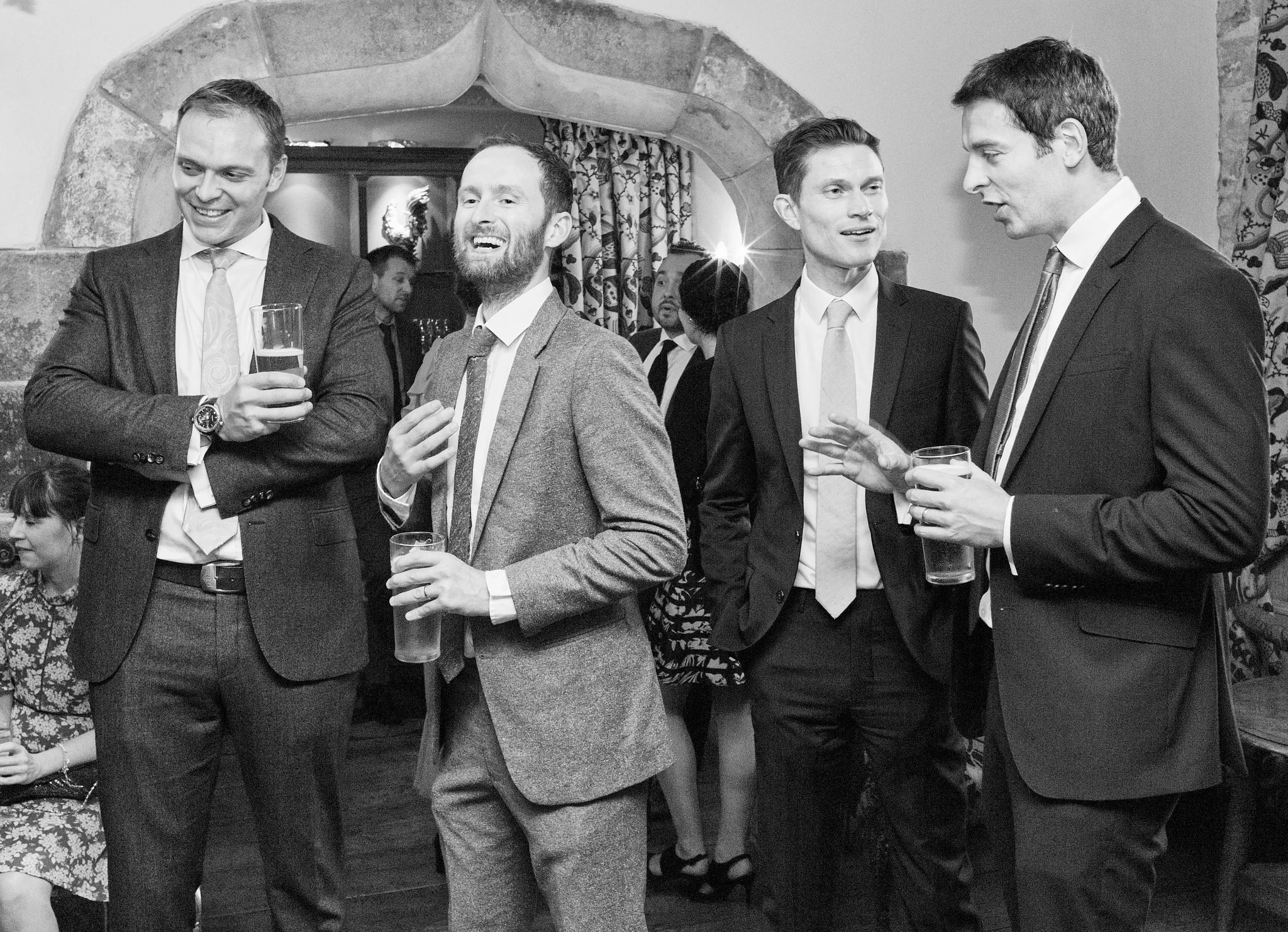 A group of four men in suits are socializing at an indoor event, holding drinks and chatting. The background features a stone archway and decorative curtains, and the photo is in black and white.