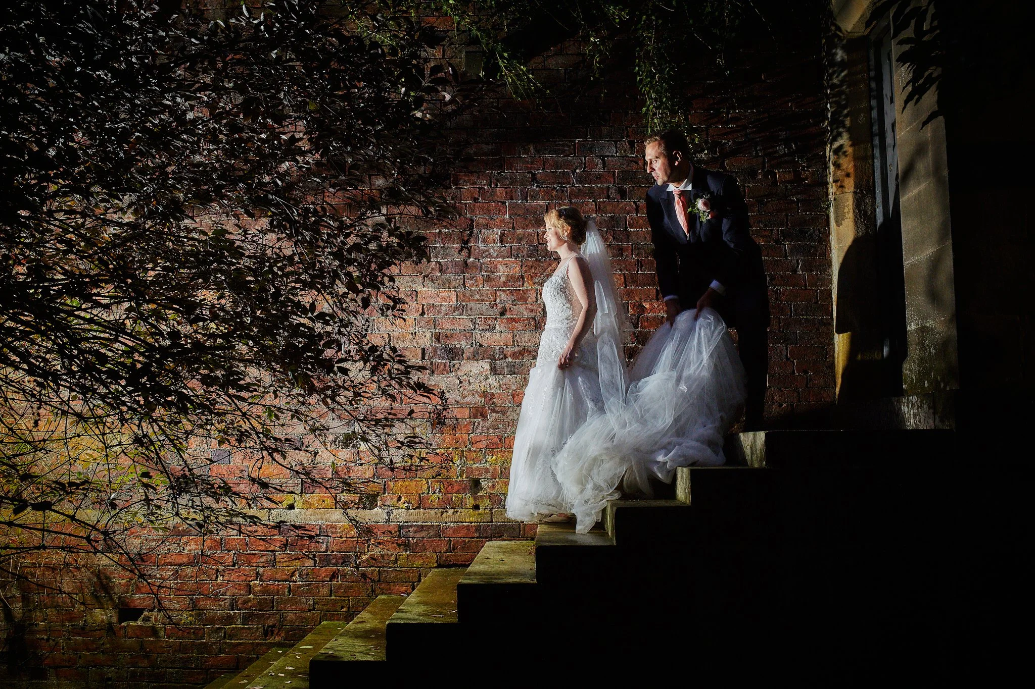 A bride in a white wedding dress and veil sitting on steps beside a groom in a dark suit with a pink tie and boutonniere, against a brick wall with leafless branches in the foreground.