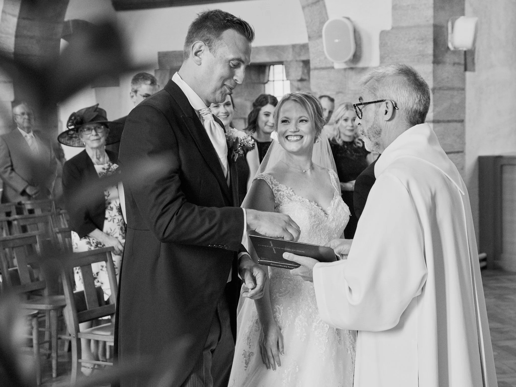 A black-and-white photo of a wedding ceremony where a groom is holding the bride's hand. The bride is smiling while looking at the officiant, who is holding a book, during the exchange of vows. Guests are seated in the background, watching the ceremo
