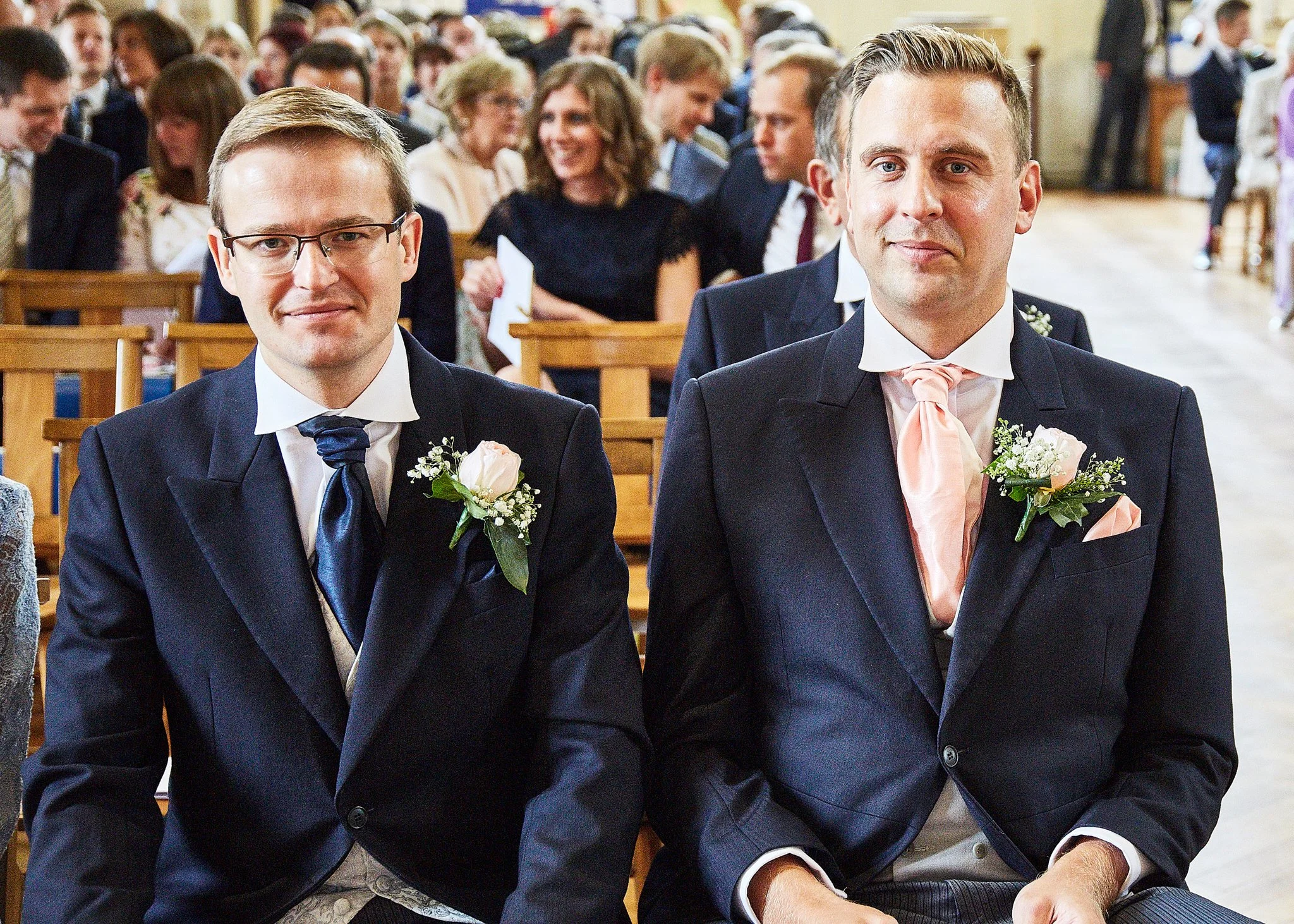 Two men dressed in suits with boutonnières sitting side by side at a wedding, surrounded by seated guests in a church.