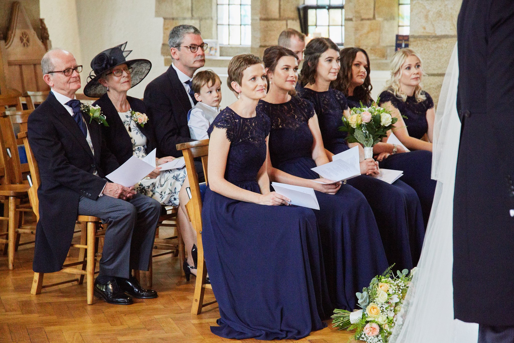 A woman in a navy blue dress is kneeling during a wedding ceremony, holding a bouquet of flowers, while other guests, including children and adults, are seated behind her, watching the ceremony.