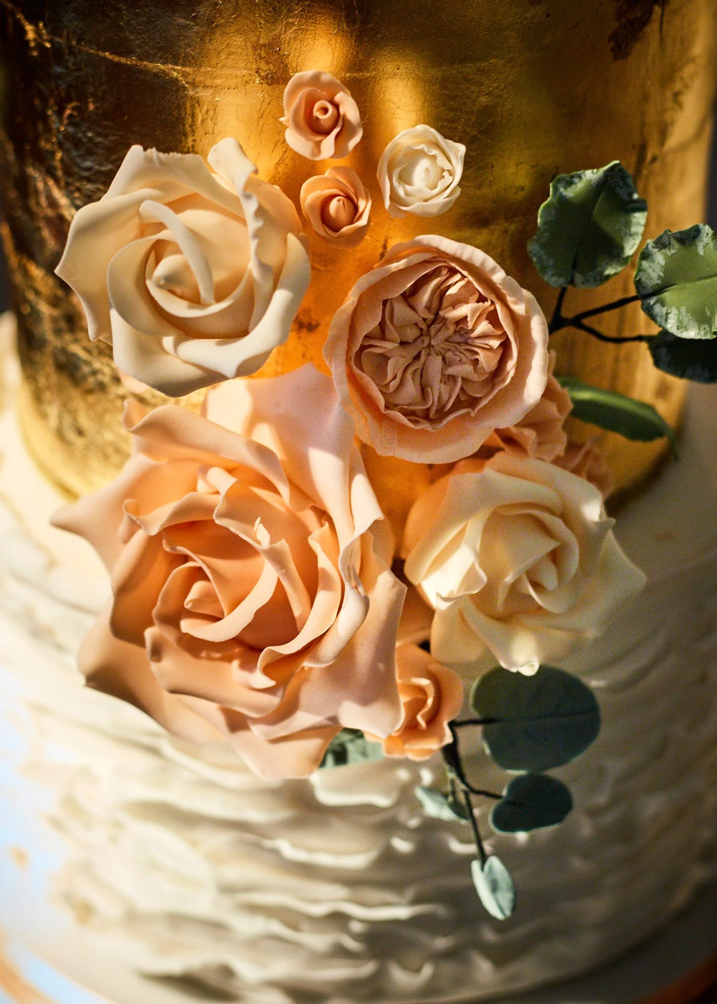 A close-up of a cake decorated with pink and white sugar roses and green leaves, with a gold and textured background.