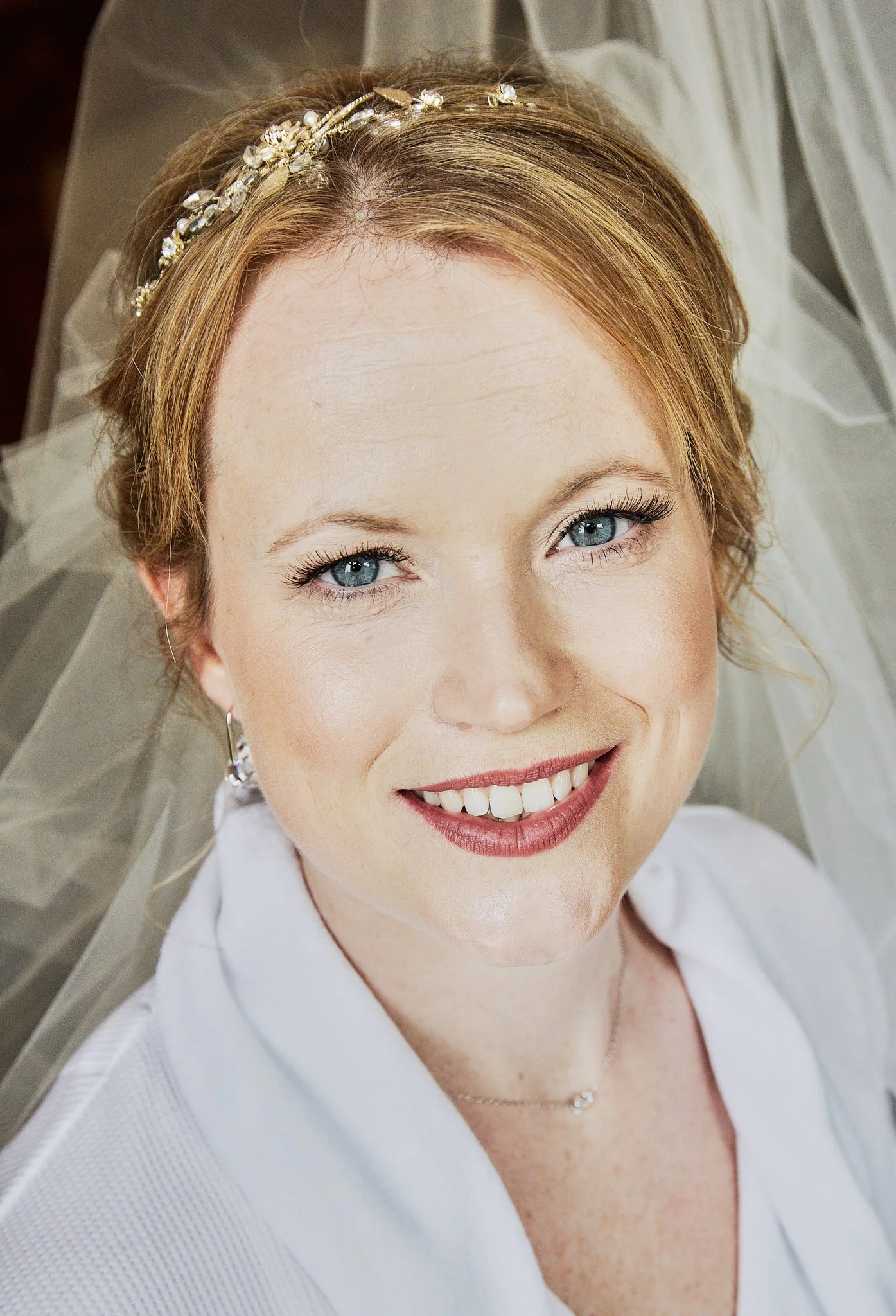 Close-up of a smiling woman with blue eyes, wearing a white blouse, a delicate necklace, and a floral headband, lying on a white surface with a sheer fabric surrounding her.
