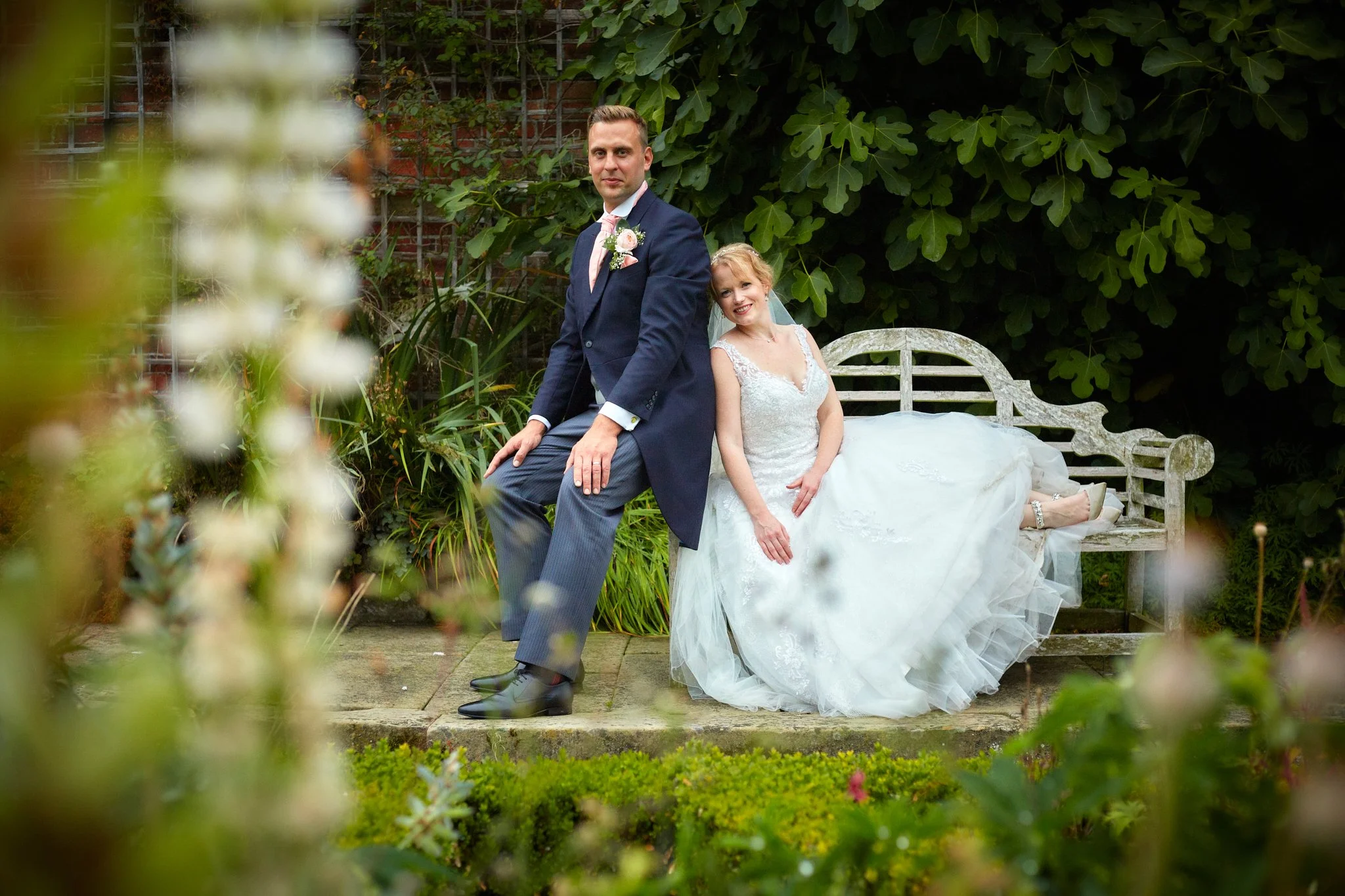 A newlywed couple sitting on a garden bench surrounded by greenery. The groom is wearing a navy suit and pink tie, and the bride is in a white wedding dress with lace details. They are smiling with a brick wall and lush foliage in the background.