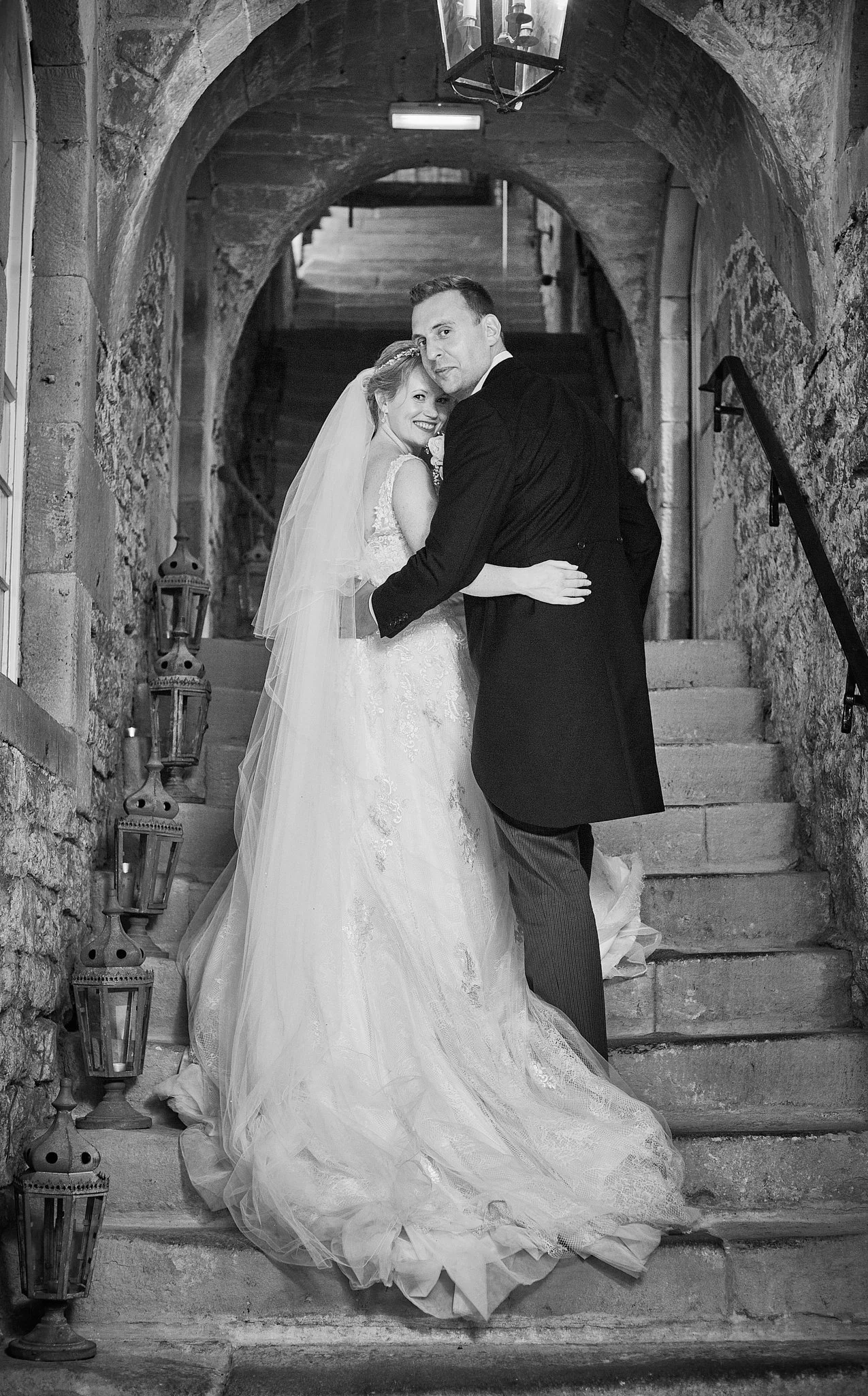 A bride and groom standing on a stone staircase in an arched stone corridor, smiling and embracing each other. The bride is in a wedding gown with a veil, and the groom is in a tuxedo. Lanterns are positioned on the stairs.