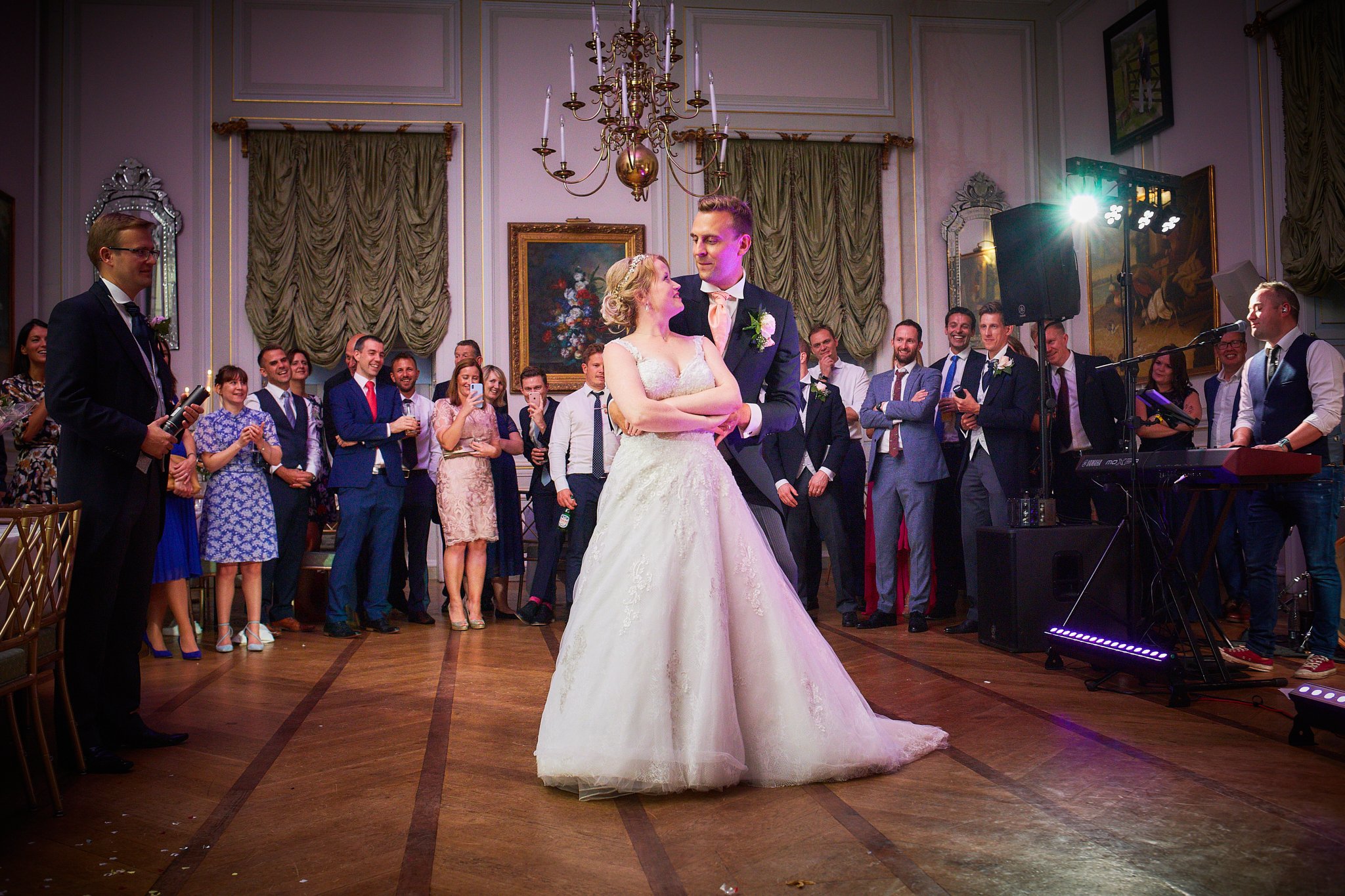 Bride and groom dancing during a wedding reception, surrounded by guests in a grand hall with ornate decorations and gold accents.