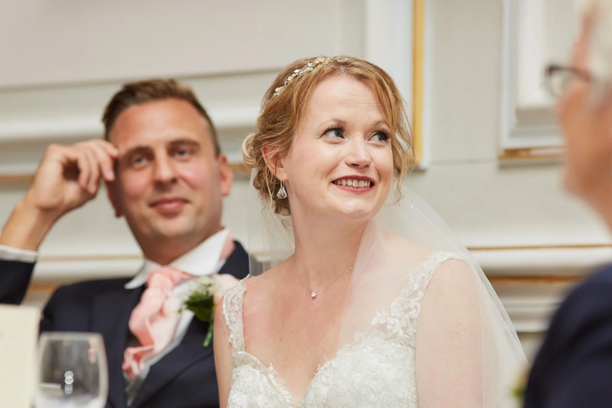 A bride and groom are at a wedding reception, sitting at the head table. The bride is smiling and looking to her right, wearing a lace wedding dress and a floral headband. The groom is sitting beside her, slightly out of focus, wearing a tuxedo and p