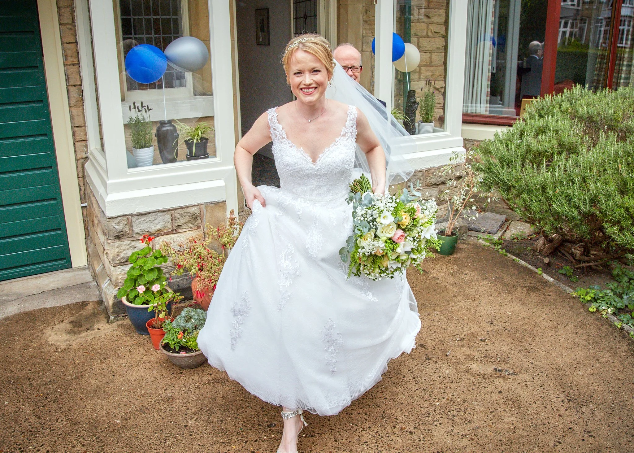 A smiling bride in a white wedding dress holding a bouquet of white and pink flowers, walking out of a house with a garden background.