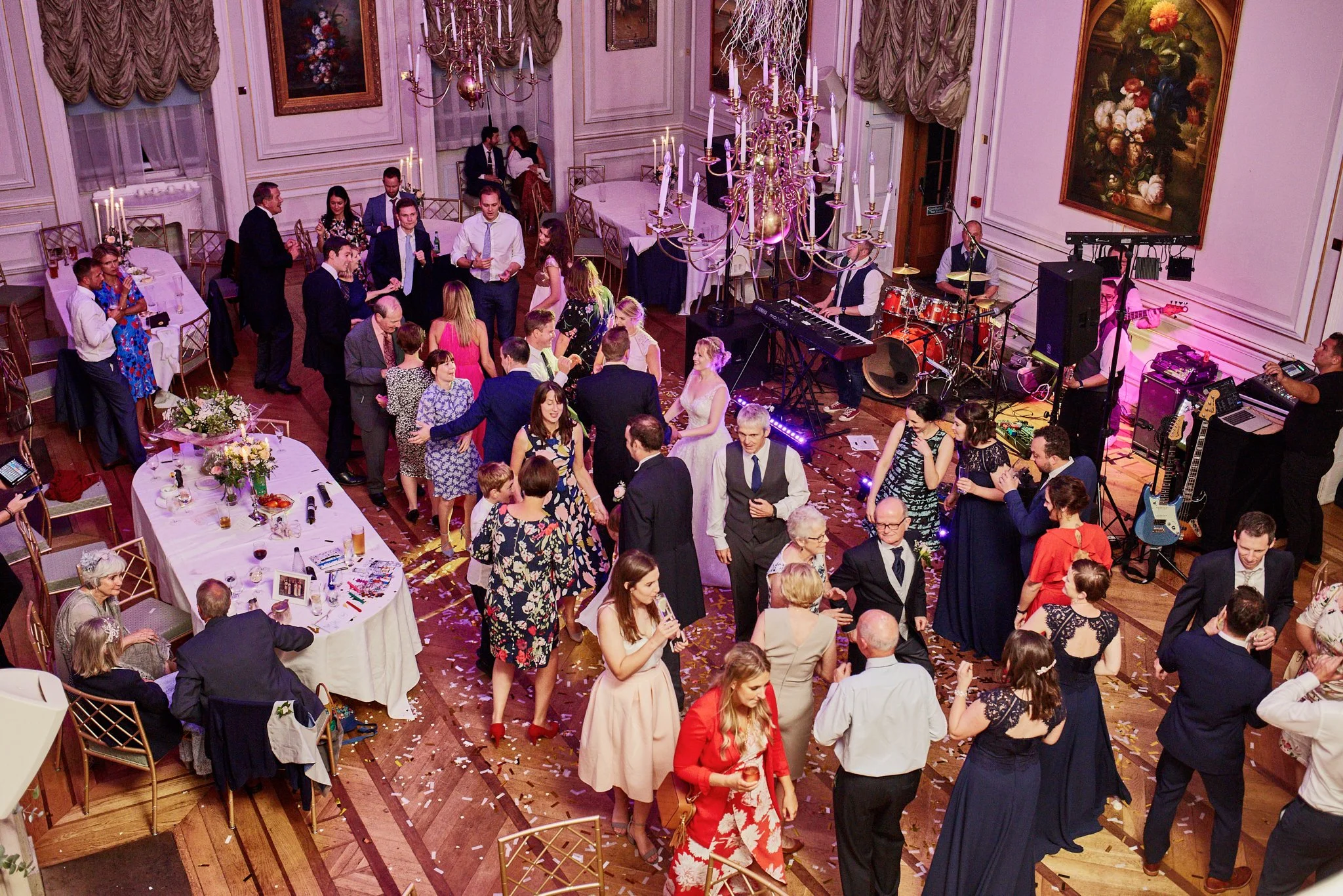 A lively indoor celebration with dancing and socializing, featuring a band setup with musical instruments, surrounded by elegantly dressed guests, floral arrangements, and ornate chandeliers in a richly decorated room.