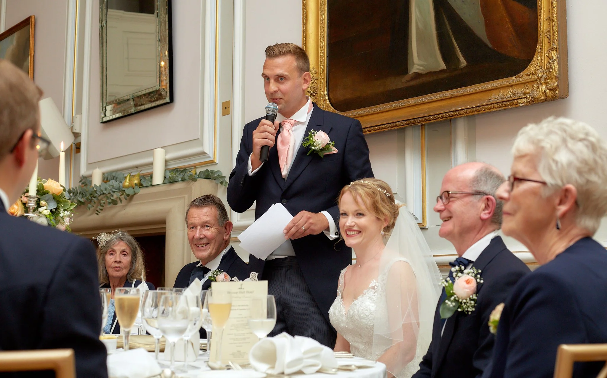 A man in a navy suit and pink tie giving a speech at a wedding reception, standing behind seated guests, including a bride in a white dress and veil, and a groom, at a decorated dining table with glasses of champagne, candles, and floral arrangements