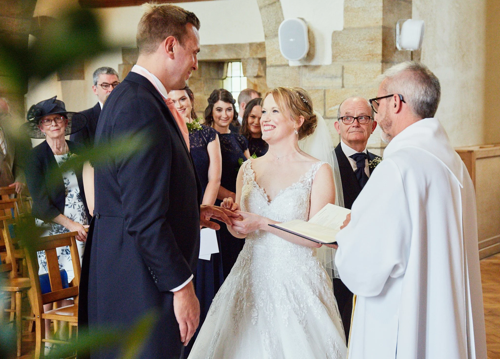 A bride and groom holding hands during their wedding ceremony in a church, with the officiant reading from a book, and guests smiling in the background.