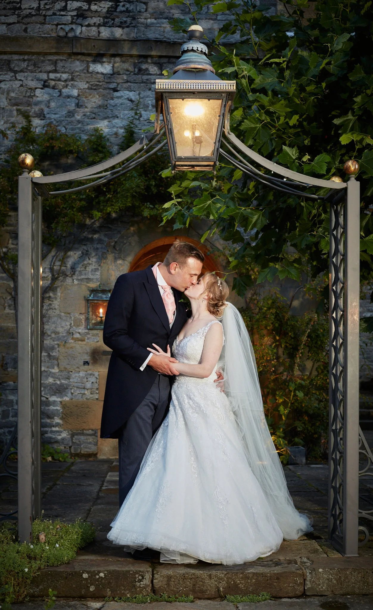 A bride and groom share a kiss under an outdoor lamp archway at dusk, with stone walls and greenery in the background.