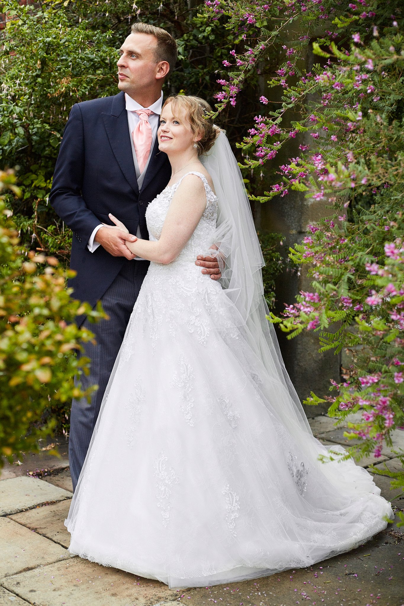 A bride and groom standing outdoors surrounded by flowering bushes. The bride is in a white wedding dress with a veil, and the groom is in a dark suit with a pink tie. They are holding hands and smiling softly, with the bride looking up and the groom