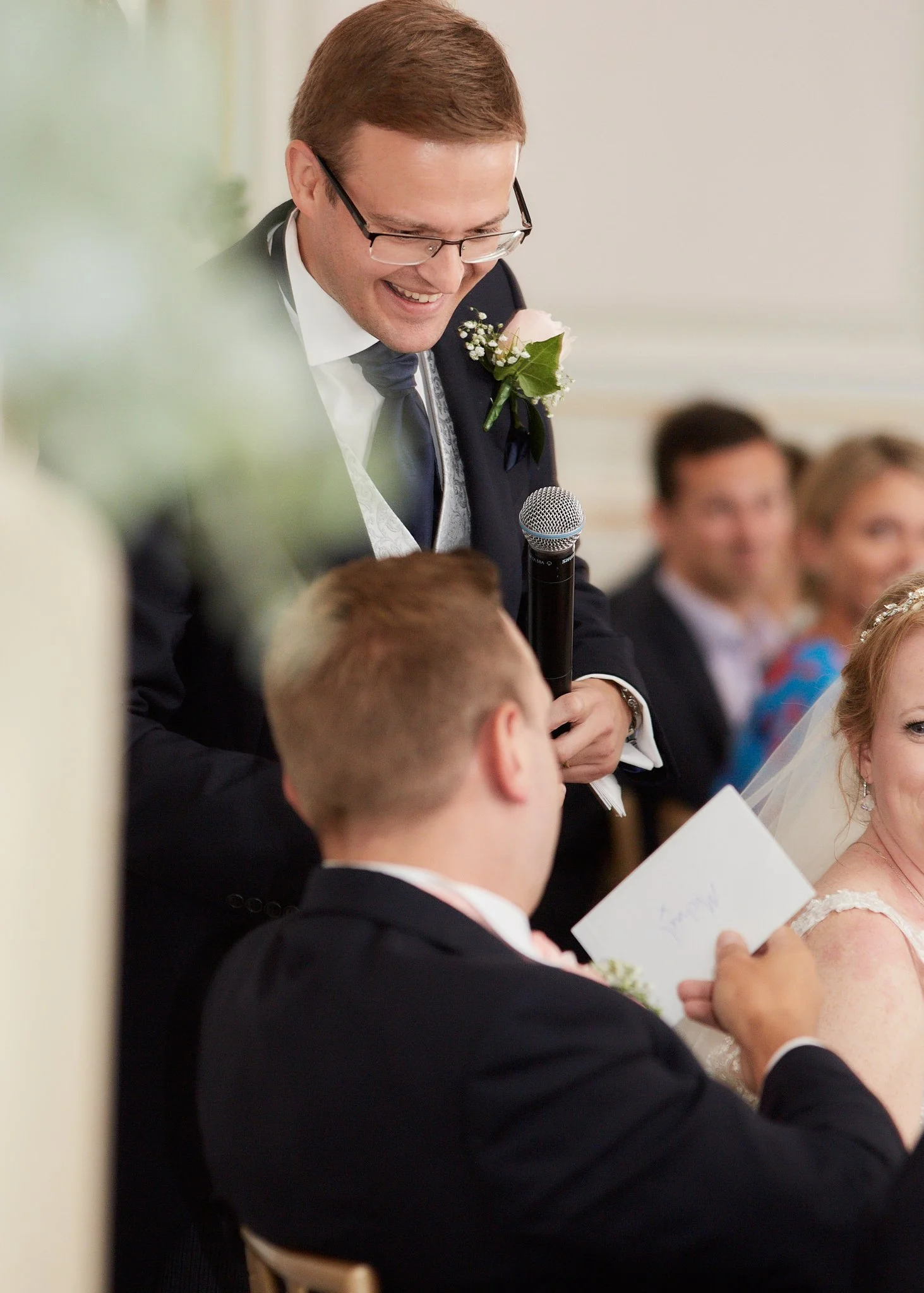 A man with glasses, wearing a suit and a boutonniere, speaking into a microphone during a wedding ceremony.
