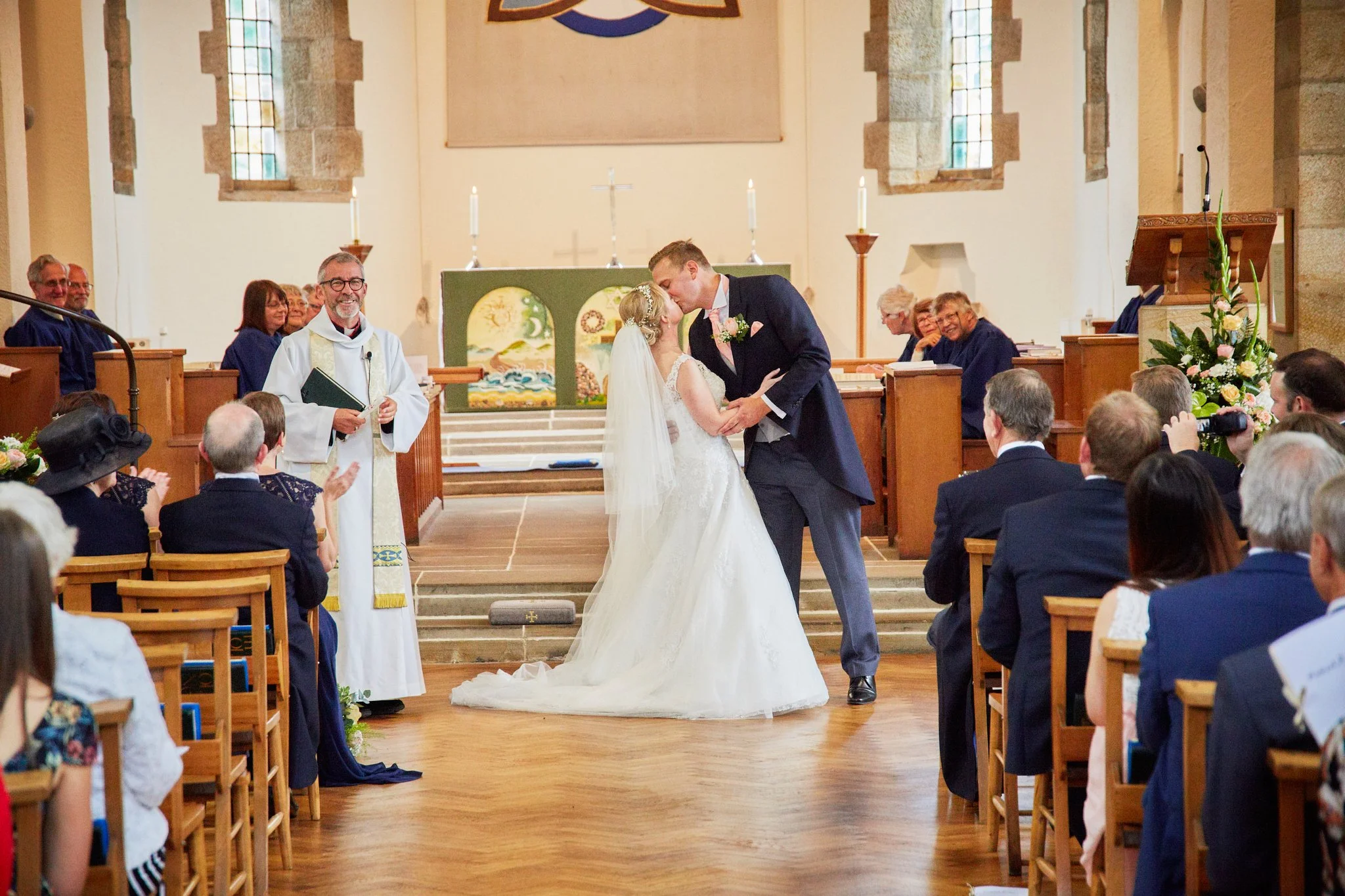 A wedding ceremony inside a church with a bride and groom kissing at the altar, surrounded by seated guests and officiant, with religious symbols and candles in the background.