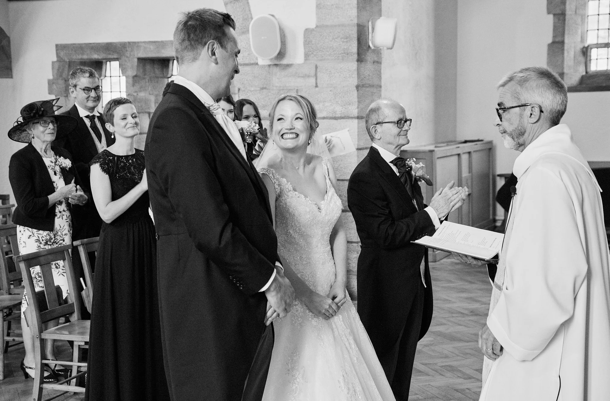 Black and white photo of a wedding ceremony in a church. The bride is smiling, holding hands with the groom, who is looking at her. The officiant stands in front of them. In the background, guests are seated and observing the ceremony.