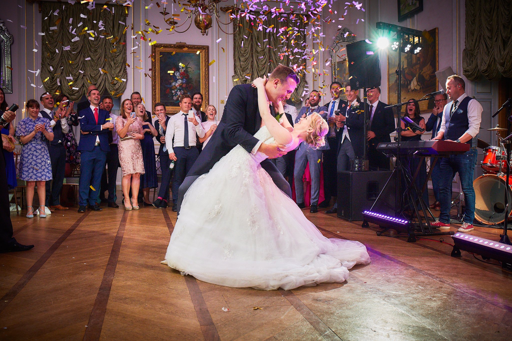 A bride and groom dancing at their wedding reception surrounded by guests. Confetti falls from the ceiling, and the guests are watching and taking photos. The scene is joyful and celebratory in a decorated ballroom.