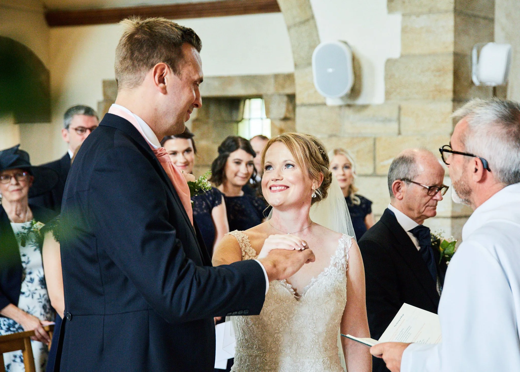 A bride and groom exchanging vows at their wedding ceremony, surrounded by friends and family in a rustic indoor setting.