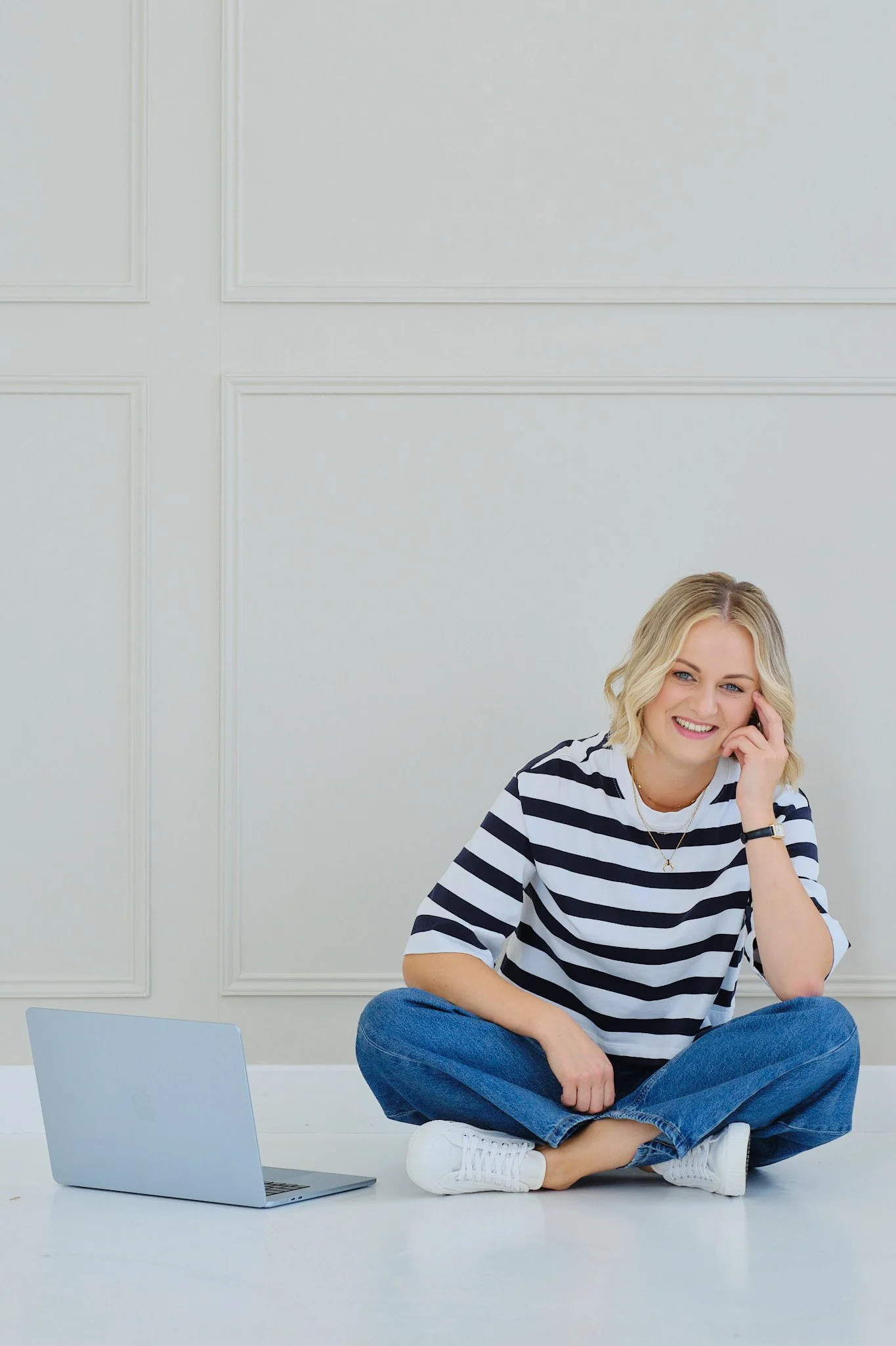 Smiling woman sitting cross-legged on the floor, talking on her phone, with a laptop and tablet nearby, in a bright room with white paneled walls.