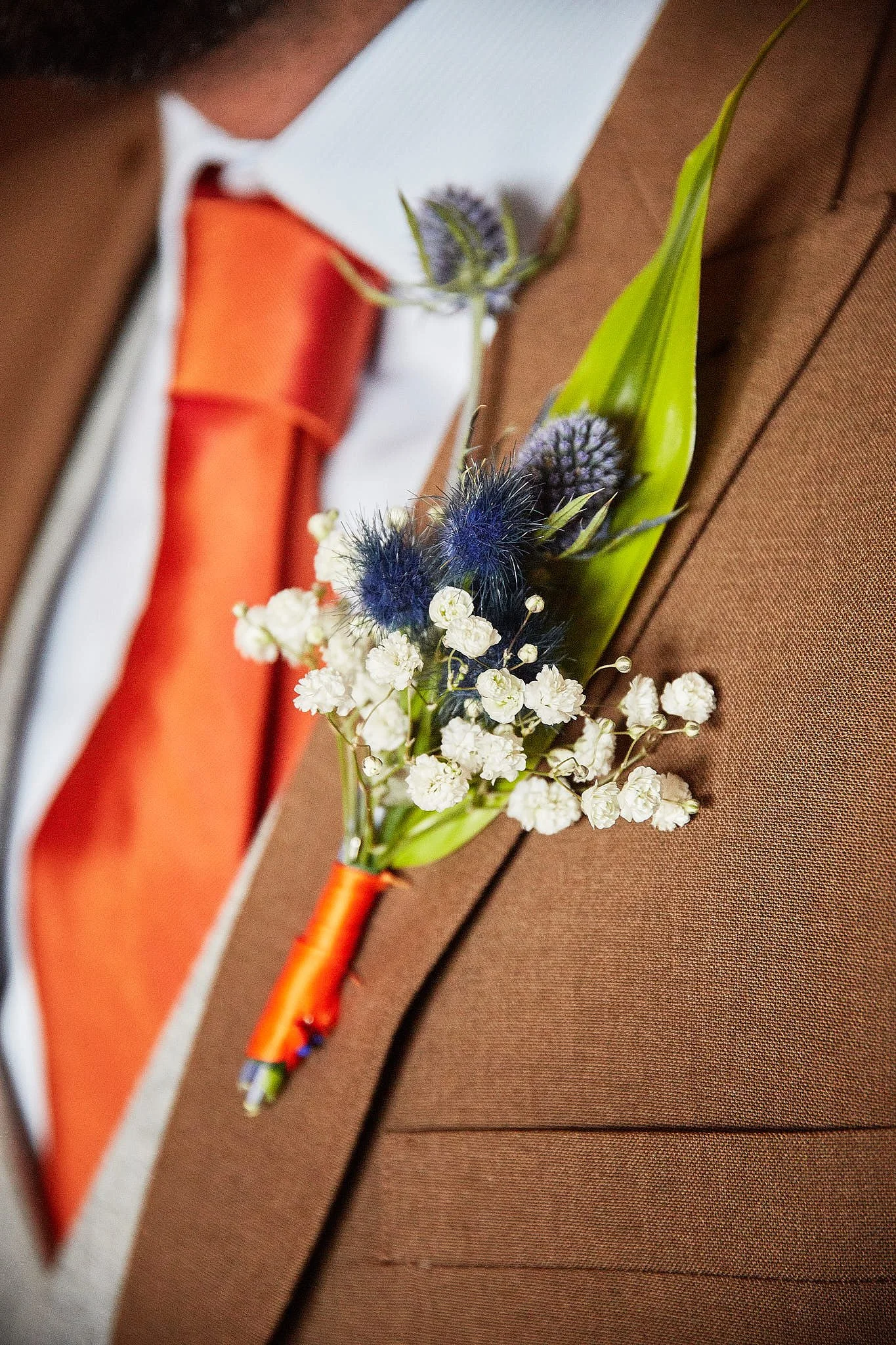 Close-up of a small boutonniere with white baby's breath flowers, purple thistle, green leaves, pinned to a brown suit jacket.