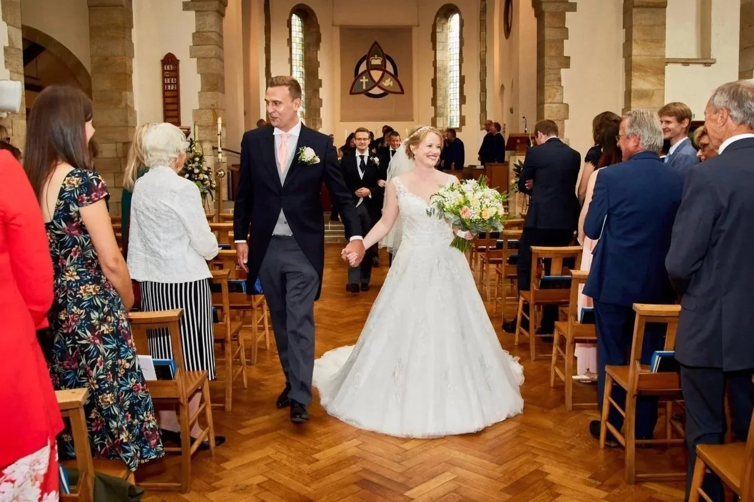 A bride and groom walking down the aisle in a church after their wedding ceremony, holding hands and smiling, with friends and family seated on both sides.