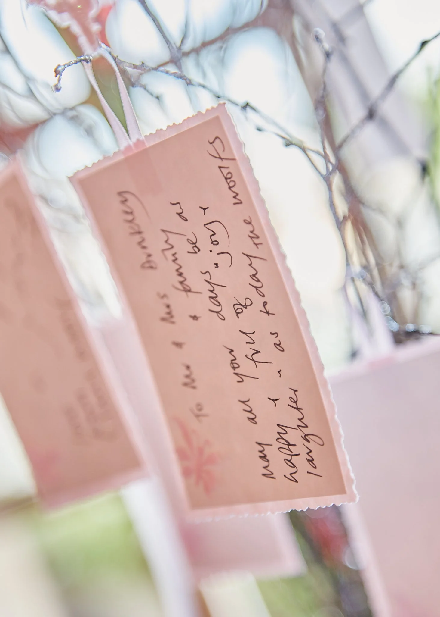 Close-up of a pink tag with handwritten text on a branch with white buds or flowers, possibly a cherry blossom.