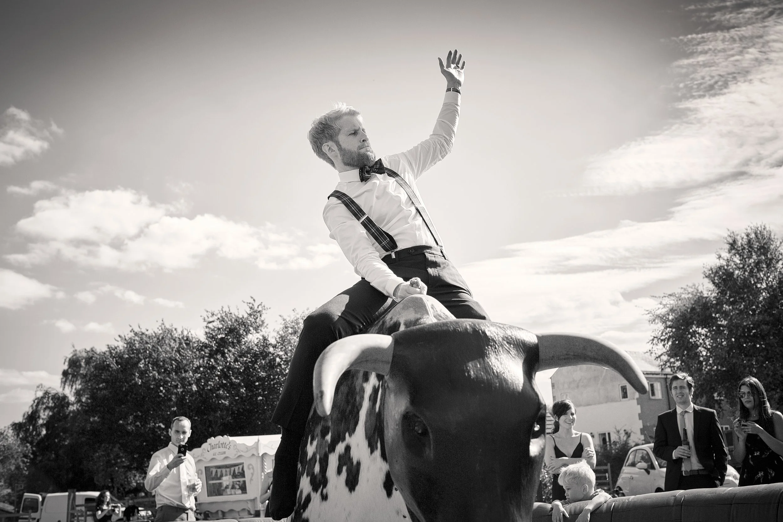 A man with a beard and bow tie is riding on a mechanical bull at an outdoor event, raising one hand in the air. Several people are watching and taking pictures in the background, with trees and buildings visible under a partly cloudy sky.