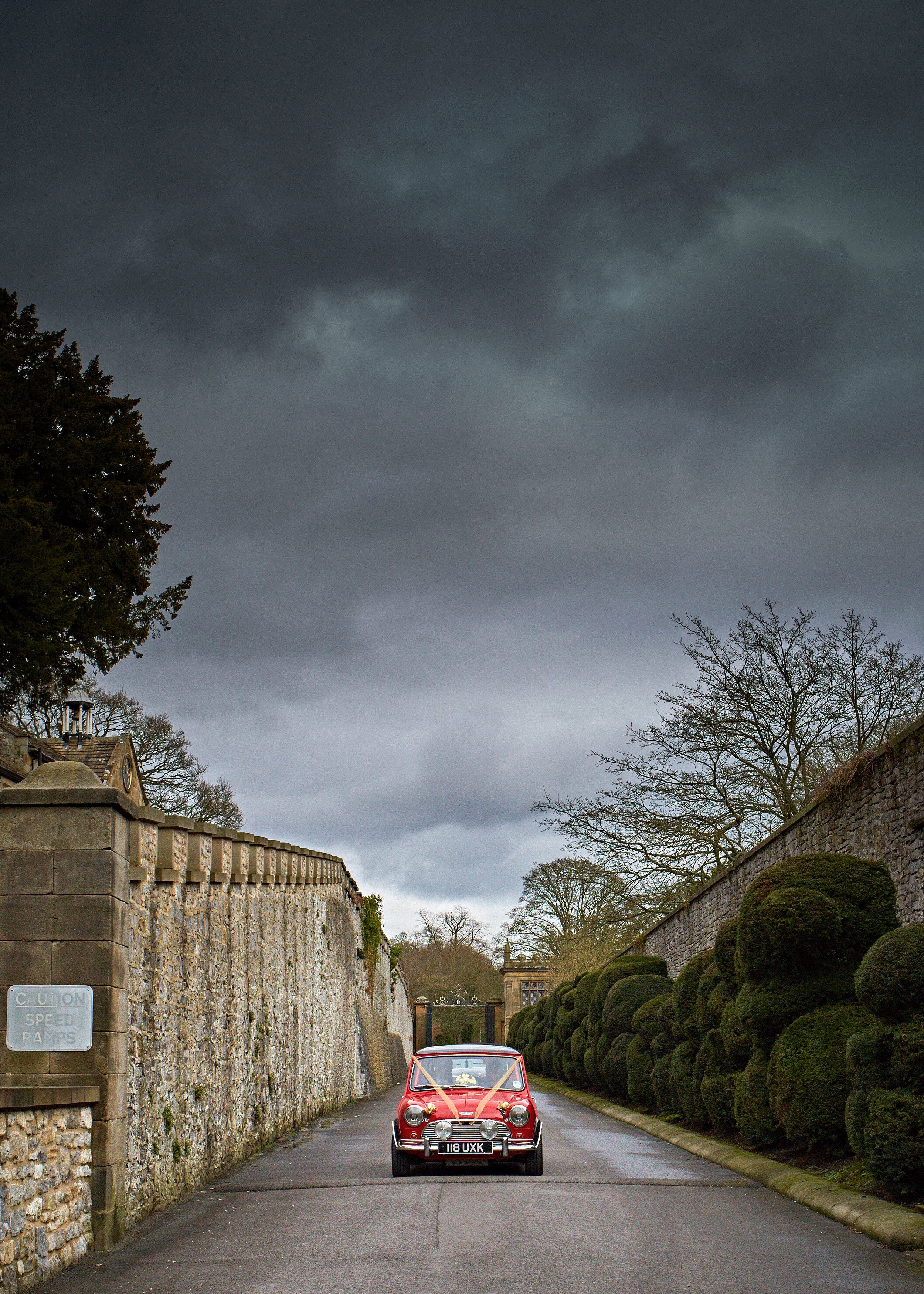 A red vintage car with ribbon decorations drives down a narrow street lined with stone walls and well-trimmed bushes, under a dark cloudy sky.