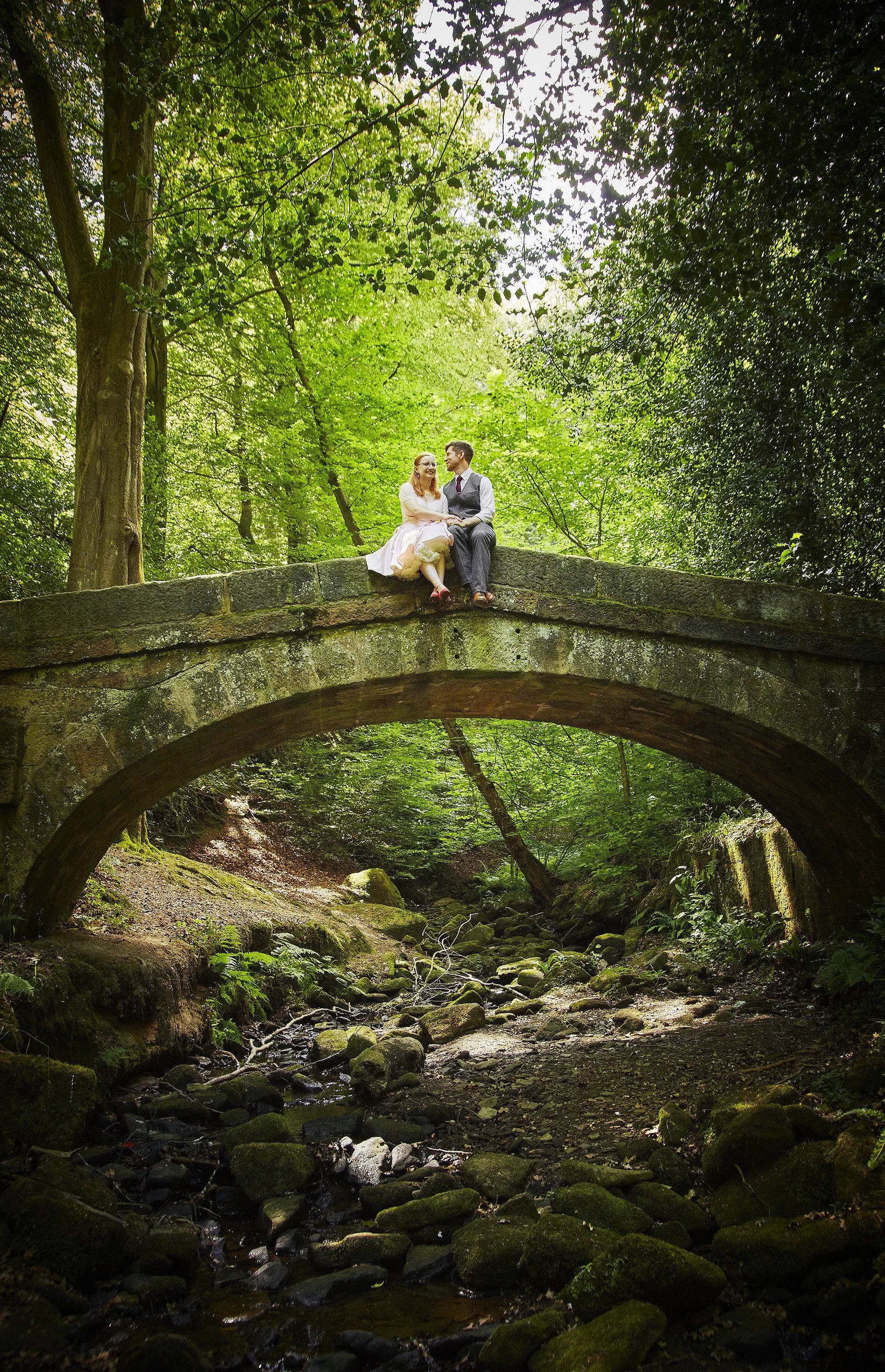 A couple sitting on a stone bridge in a lush green forest, with a small stream running under the bridge.