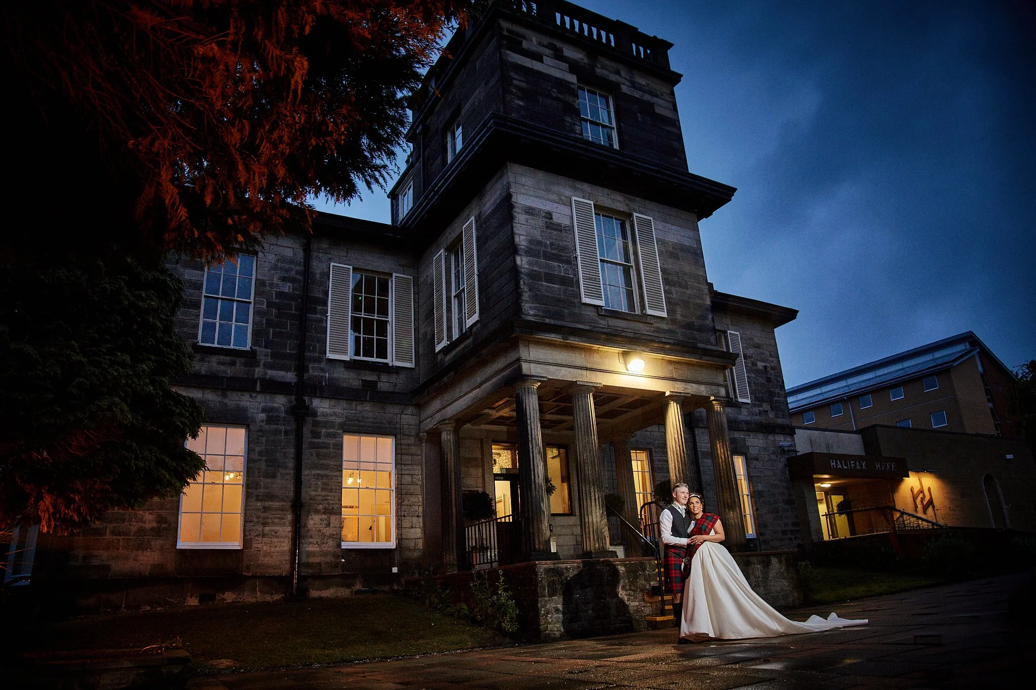 A couple dressed in wedding attire standing outside a historic building at night. The bride is wearing a white gown with a long train, and the groom is in a gray vest and tie. The building has large windows with warm light glowing from inside, stone 