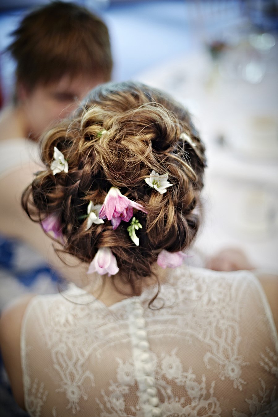 Brunette woman with her hair styled in an updo decorated with white and pink flowers, wearing a lace dress, at a formal event.